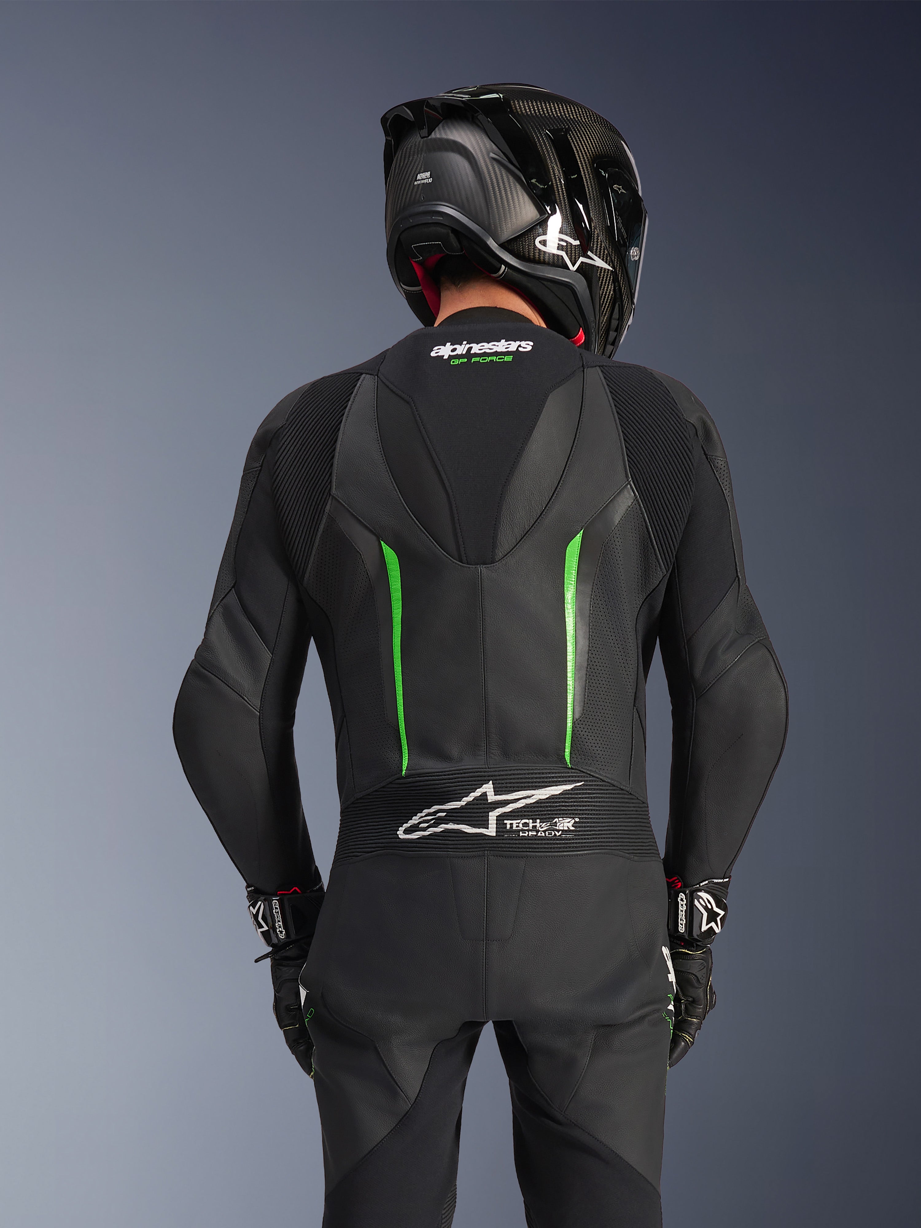Widok z tyłu motocyklisty w skórzanym kombinezonie Alpinestars GP Force V2 w kolorze Black Green, z aerodynamicznym garbem, oznaczeniem Tech-Air Ready na harmonijkowym panelu w dolnej części pleców i konstrukcją z perforowanej skóry, w czarnym wyścigowym kasku z włókna węglowego.