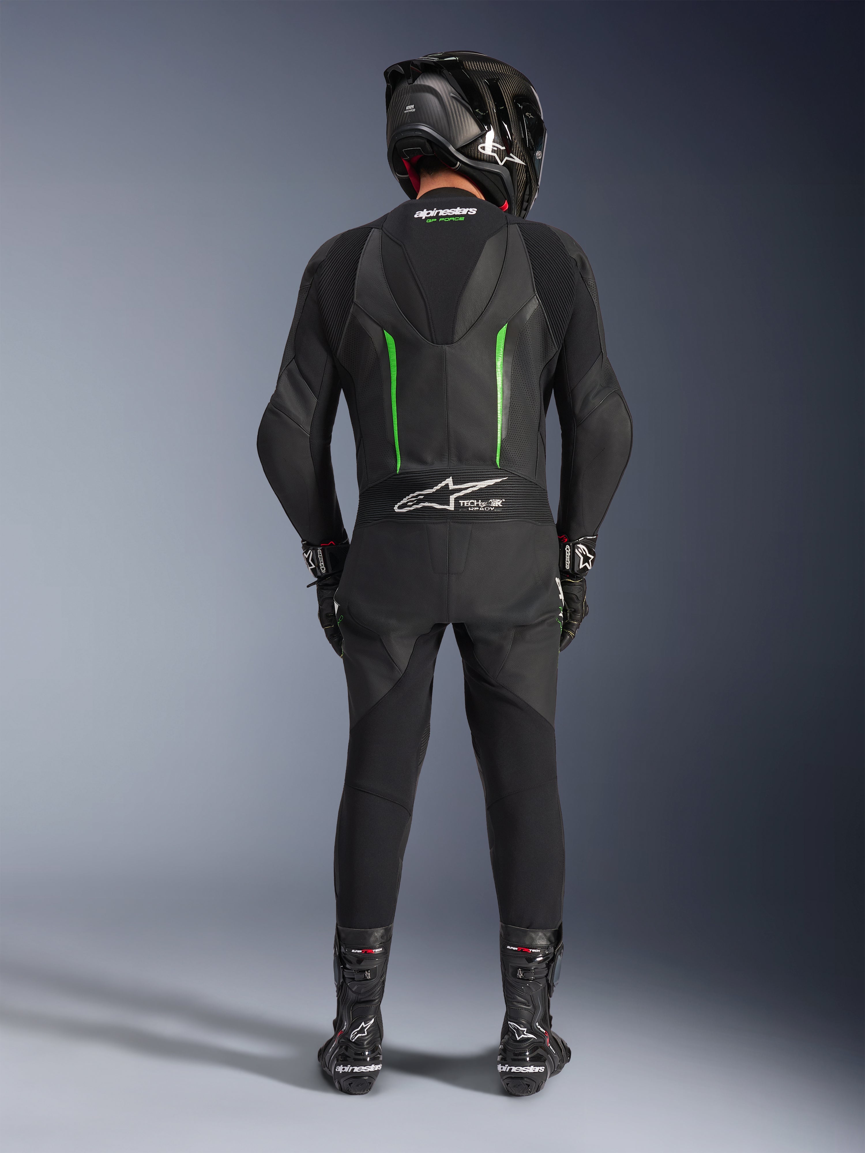 Motocyklista widoczny od tyłu w jednoczęściowym skórzanym kombinezonie Alpinestars GP Force V2 w kolorze czarno-zielonym, czarny kask z włókna węglowego, czarne rękawice wyścigowe i czarne buty, aerodynamiczny garb oraz branding Tech-Air Ready.
