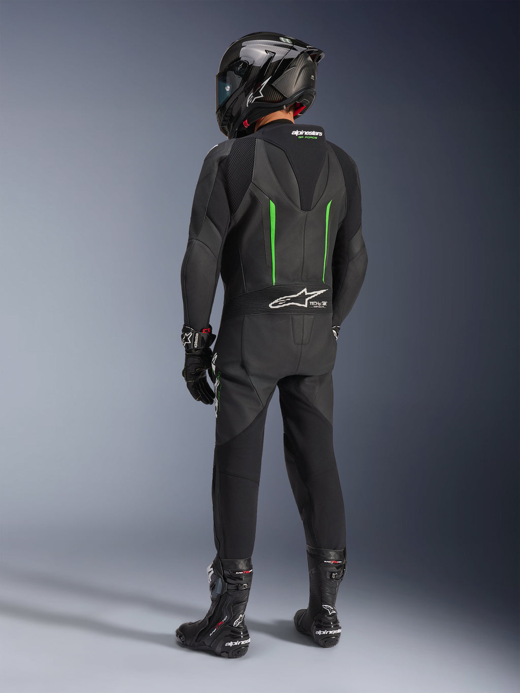 Motocyklista widoczny od tyłu w jednoczęściowym skórzanym kombinezonie Alpinestars GP Force V2, czarny z zielonymi akcentami, aerodynamiczny garb, czarny kask z włókna węglowego, czarne buty wyścigowe i rękawice, wysokiej klasy wyczynowa odzież motocyklowa