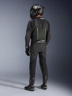 Motocyklista widoczny od tyłu w jednoczęściowym skórzanym kombinezonie Alpinestars GP Force V2, czarny z zielonymi akcentami, aerodynamiczny garb, czarny kask z włókna węglowego, czarne buty wyścigowe i rękawice, wysokiej klasy wyczynowa odzież motocyklowa