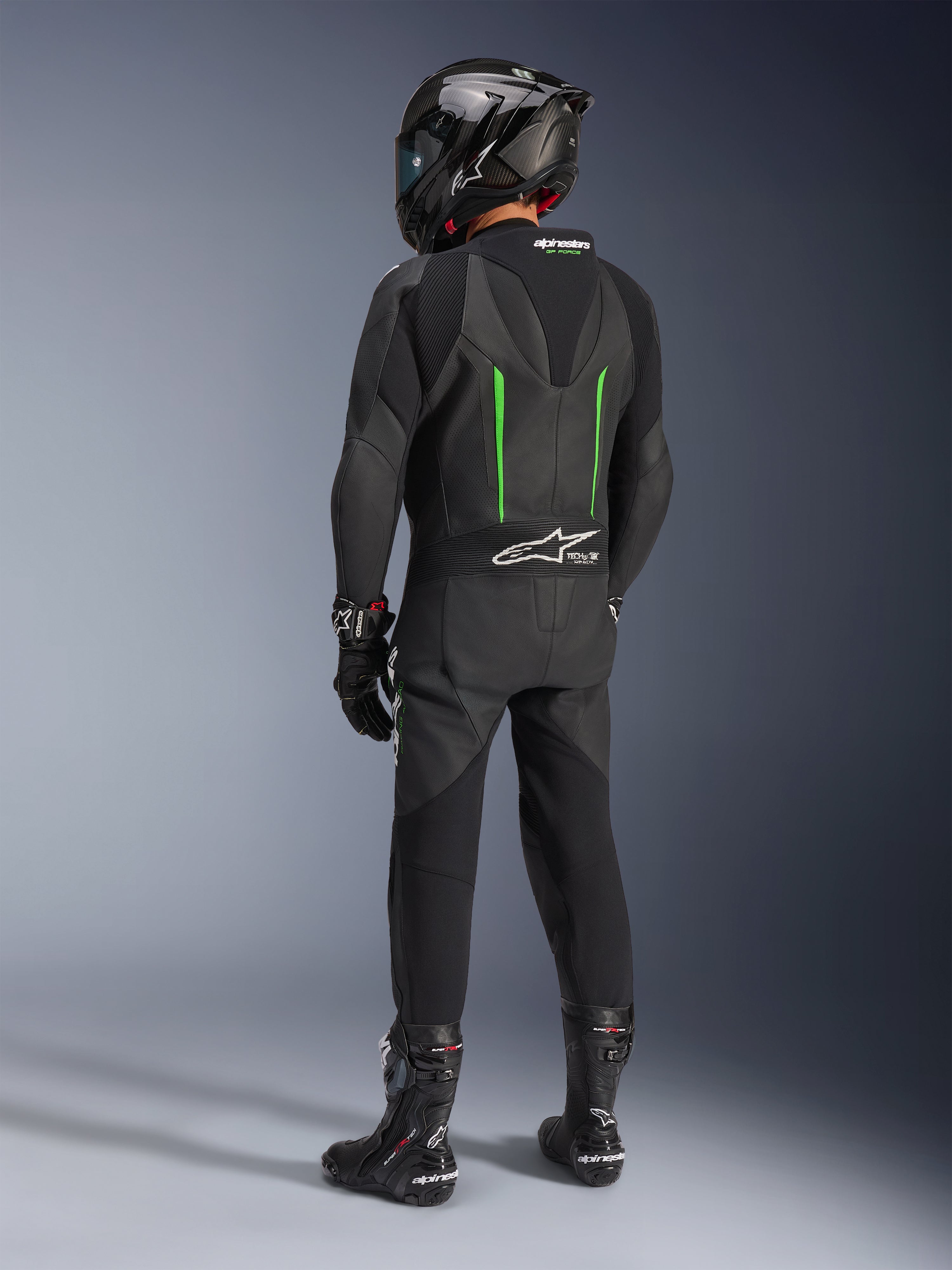 Motocyklista widoczny od tyłu w jednoczęściowym skórzanym kombinezonie Alpinestars GP Force V2, czarny z zielonymi akcentami, aerodynamiczny garb, czarny kask z włókna węglowego, czarne buty wyścigowe i rękawice, wysokiej klasy wyczynowa odzież motocyklowa