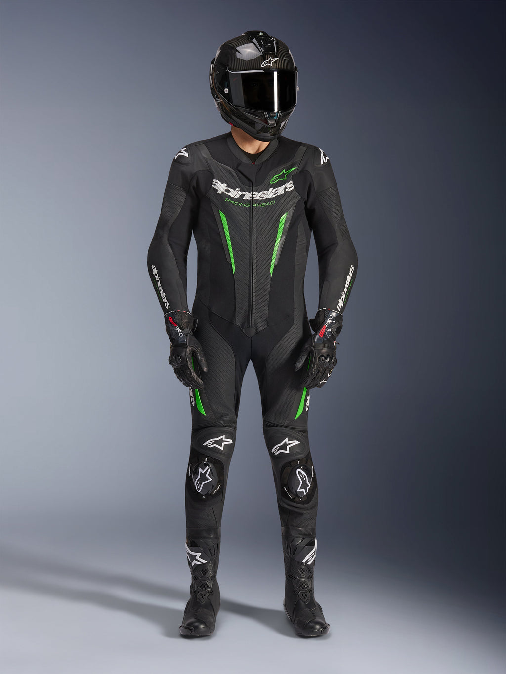 Motocyklista wyścigowy ubrany w jednoczęściowy kombinezon skórzany Alpinestars GP Force V2 w kolorze czarno-zielonym, w zestawie z kaskiem z włókna węglowego i rękawicami wyścigowymi, z perforowanymi panelami skórzanymi i białymi logo Alpinestars, profesjonalny sprzęt do jazdy torowej i sportowej
