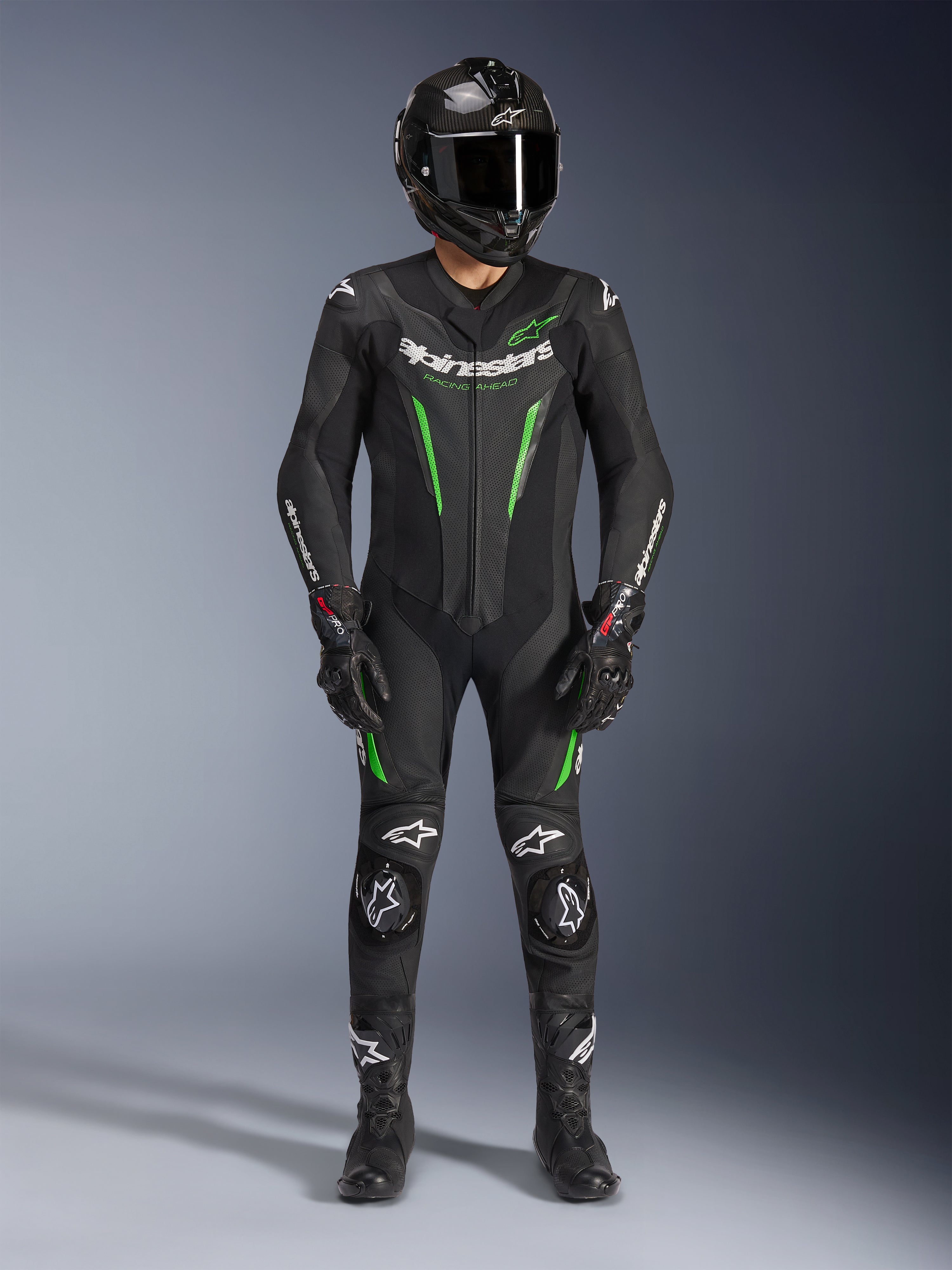 Motocyklista wyścigowy ubrany w jednoczęściowy kombinezon skórzany Alpinestars GP Force V2 w kolorze czarno-zielonym, w zestawie z kaskiem z włókna węglowego i rękawicami wyścigowymi, z perforowanymi panelami skórzanymi i białymi logo Alpinestars, profesjonalny sprzęt do jazdy torowej i sportowej