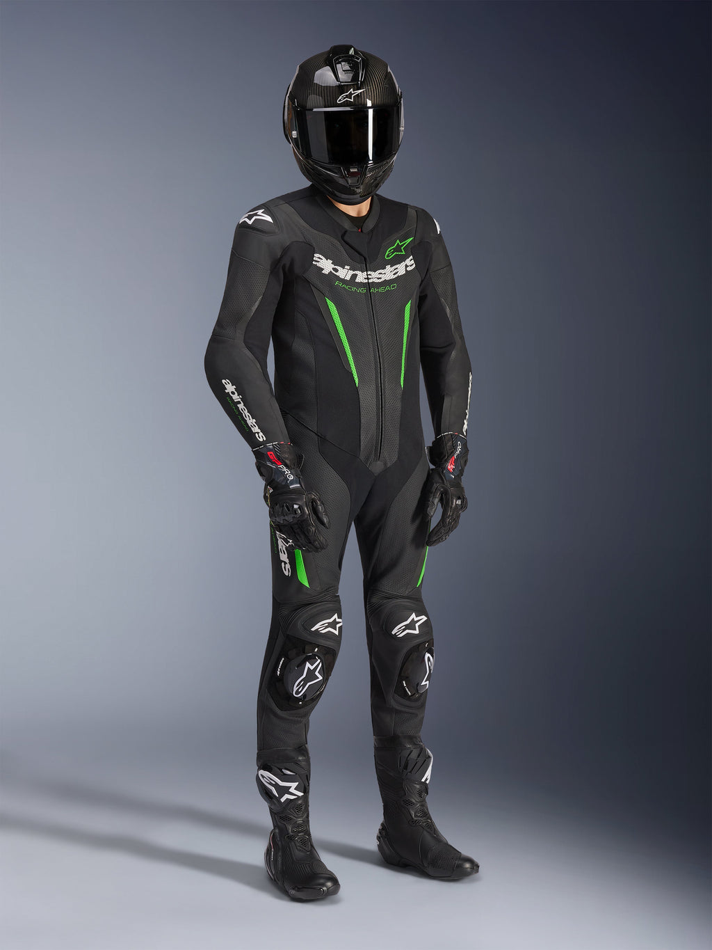 Jednoczęściowy skórzany kombinezon Alpinestars GP Force V2, czarno-zielony, noszony z dopasowanym czarnym kaskiem, rękawicami wyścigowymi i butami, z jasnozielonymi akcentami i wyraźnym białym brandingiem Alpinestars, profesjonalna odzież do jazdy torowej i sportowej