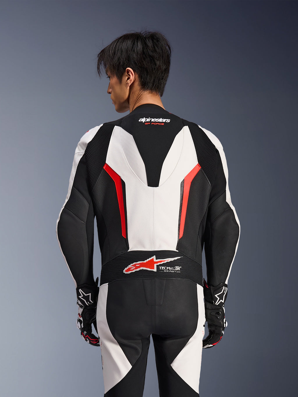 Alpinestars GP Force V2 Leather Suit 1Piece, biało-czarno-czerwony fluo, widok z tyłu na modelu, aerodynamiczny garb, perforowane panele skórzane, logo Tech-Air Ready w dolnej części pleców, profesjonalna odzież wyścigowa z czarno-białymi rękawicami