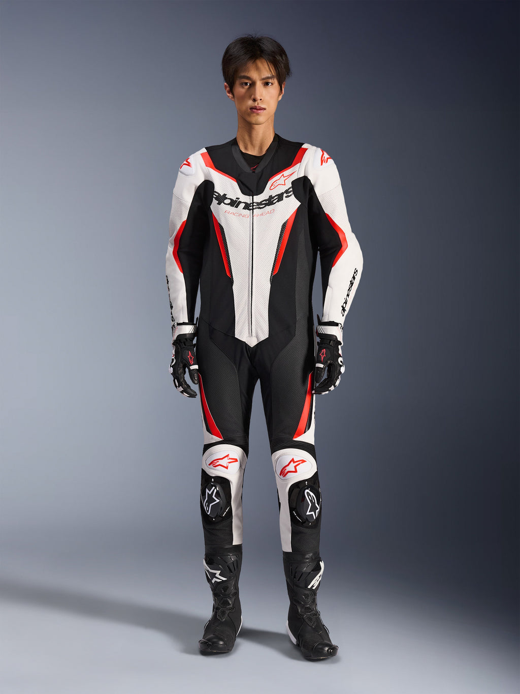 Alpinestars GP Force V2 Leather Suit - 1Piece, biało-czarno-czerwony fluorescencyjny, jednoczęściowy skórzany kombinezon motocyklowy, widok z przodu na modelu, z perforowanymi panelami i czerwonymi akcentami, wyraźne logo Alpinestars na klatce piersiowej, w stylizacji z profesjonalnymi butami i rękawicami wyścigowymi