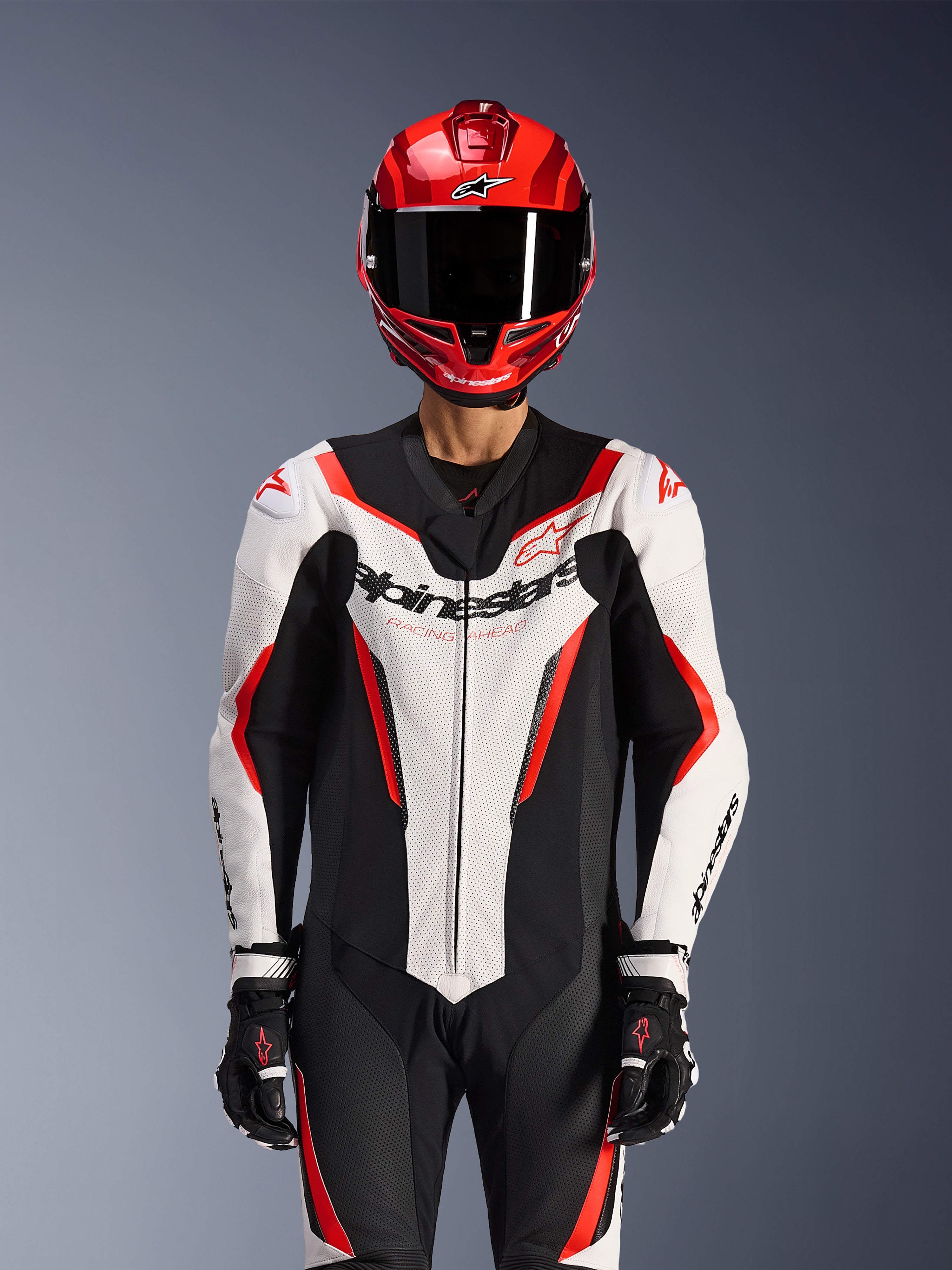 Kombinezon skórzany Alpinestars GP Force V2 - jednoczęściowy, biało-czarno-czerwony (fluo), noszony z czerwonym kaskiem integralnym i czarnymi rękawicami wyścigowymi, wyposażony w perforowane panele skórzane i wyraźne logo Alpinestars na klatce piersiowej, profesjonalna odzież wyczynowa do wyścigów motocyklowych