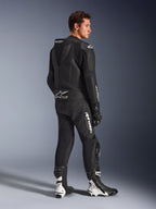 Męski model w dwuczęściowym skórzanym kombinezonie Alpinestars GP Force V2 w kolorze czarnym z białymi logotypami, widok z tyłu z profilu ukazujący aerodynamiczny garb i branding Tech-Air Ready, w zestawie z czarno-białymi butami i rękawicami wyścigowymi, profesjonalna sportowa odzież motocyklowa.