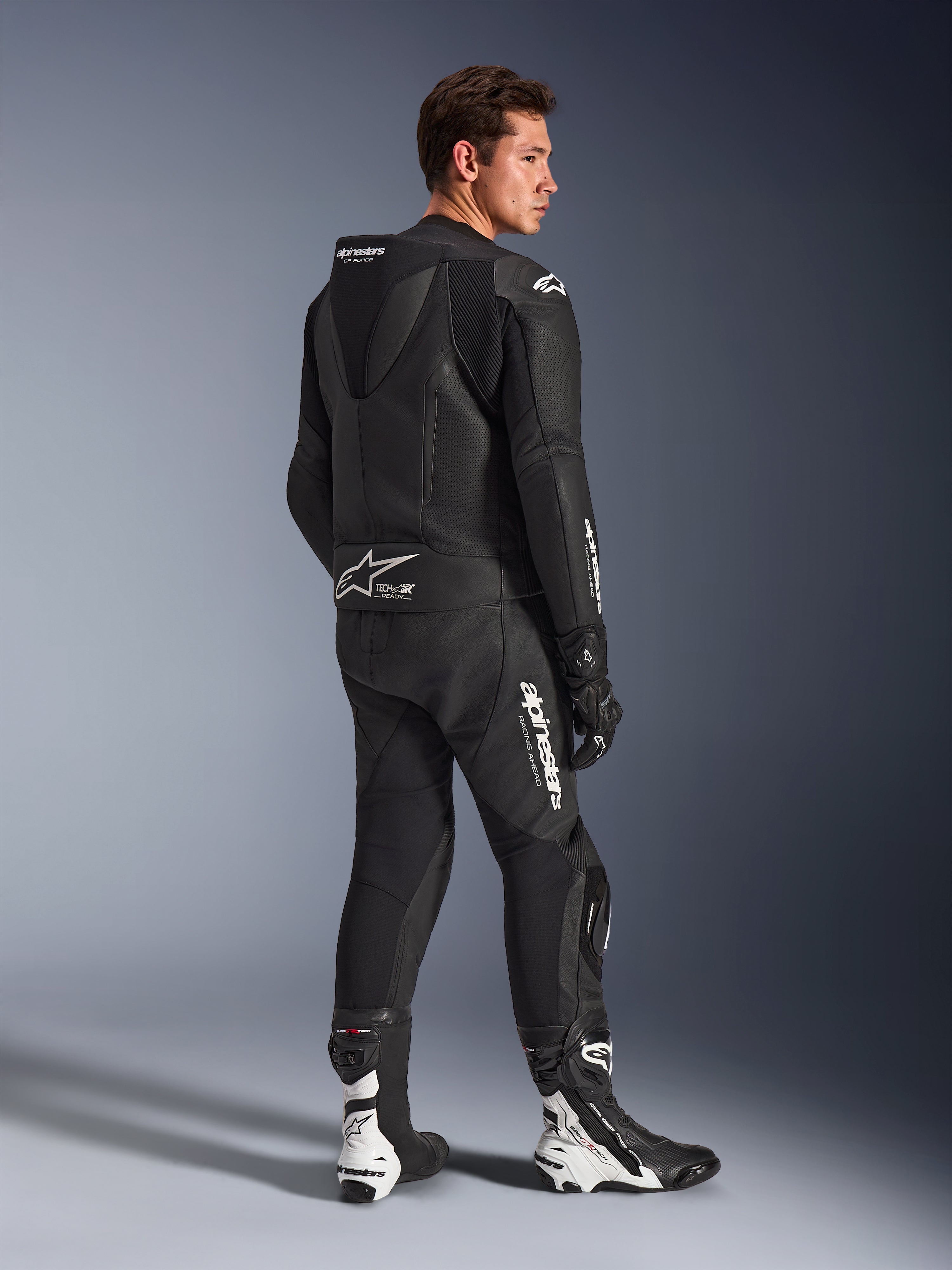 Męski model w dwuczęściowym skórzanym kombinezonie Alpinestars GP Force V2 w kolorze czarnym z białymi logotypami, widok z tyłu z profilu ukazujący aerodynamiczny garb i branding Tech-Air Ready, w zestawie z czarno-białymi butami i rękawicami wyścigowymi, profesjonalna sportowa odzież motocyklowa.