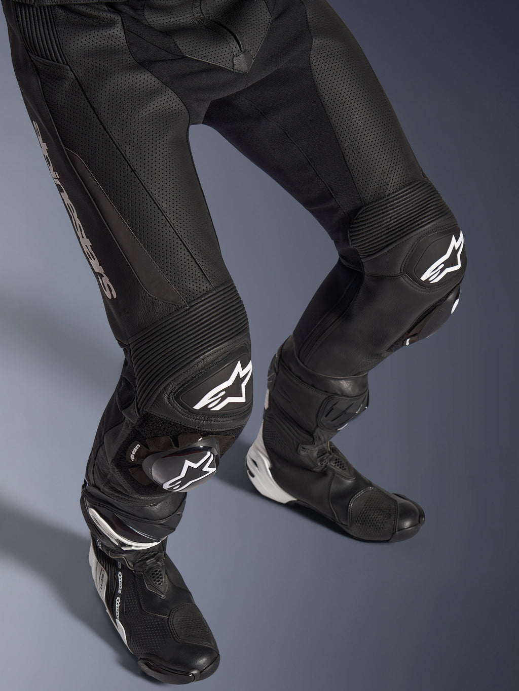Dwuczęściowy skórzany kombinezon Alpinestars GP Force V2, zbliżenie na dolną część ciała, w tym spodnie i buty wyścigowe, czarny z białym logo, cechujący się perforowaną skórzaną konstrukcją, harmonijkowymi panelami elastycznymi na kolanach i ochronnymi sliderami, profesjonalna odzież do jazdy sportowej.