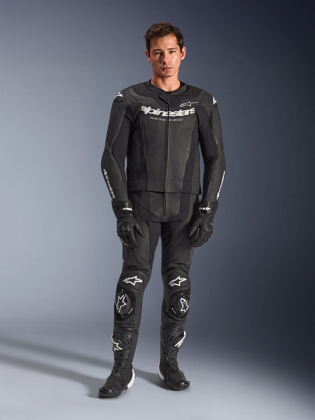 Alpinestars GP Force V2 Leather Suit 2-Piece, dwuczęściowy kombinezon skórzany, czarny z białymi logo, pełny motocyklowy kombinezon wyścigowy, wykonany z wysokiej jakości skóry z wzmocnionymi sliderami na kolanach i perforowanymi panelami, z czarnymi rękawicami i butami, widok całej sylwetki w studio