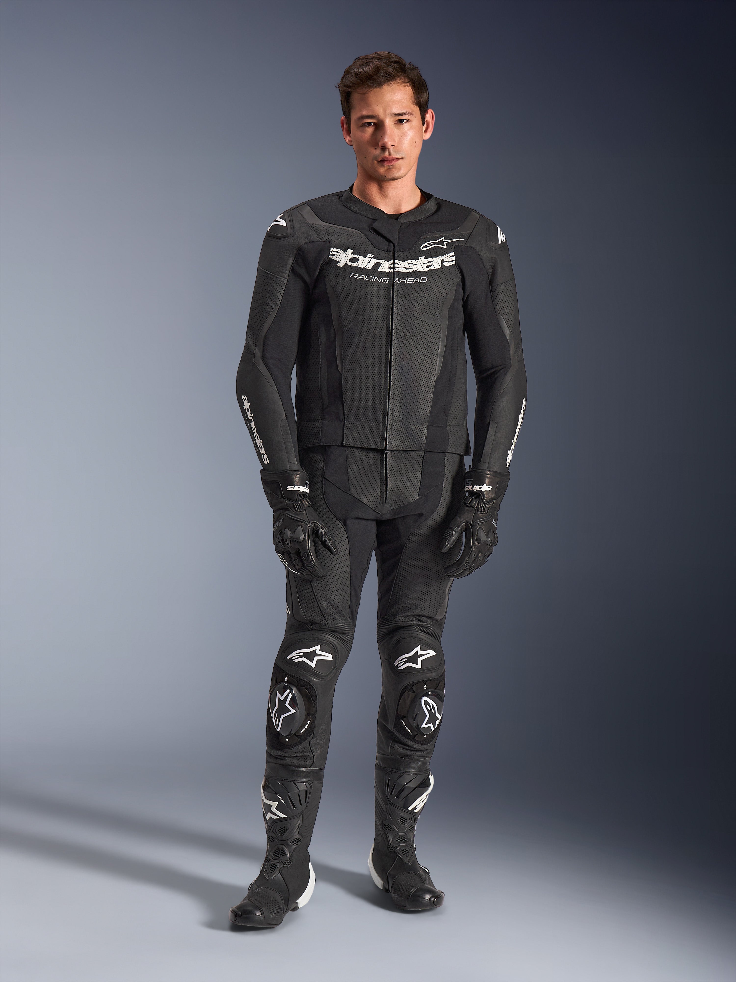 Alpinestars GP Force V2 Leather Suit 2-Piece, dwuczęściowy kombinezon skórzany, czarny z białymi logo, pełny motocyklowy kombinezon wyścigowy, wykonany z wysokiej jakości skóry z wzmocnionymi sliderami na kolanach i perforowanymi panelami, z czarnymi rękawicami i butami, widok całej sylwetki w studio