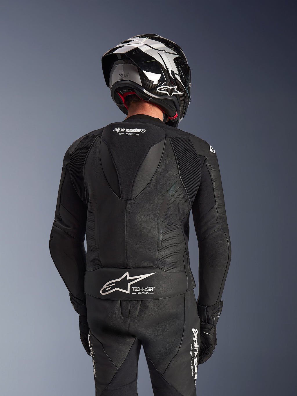 Dwuczęściowy skórzany kombinezon Alpinestars GP Force V2, czarny, widok z tyłu na motocyklistę prezentujący aerodynamiczny garb, perforowane panele skórzane oraz harmonijkowe strefy elastyczne na ramionach, z logo Tech-Air Ready w dolnej części pleców, noszony z pasującym kaskiem wyścigowym i rękawicami.