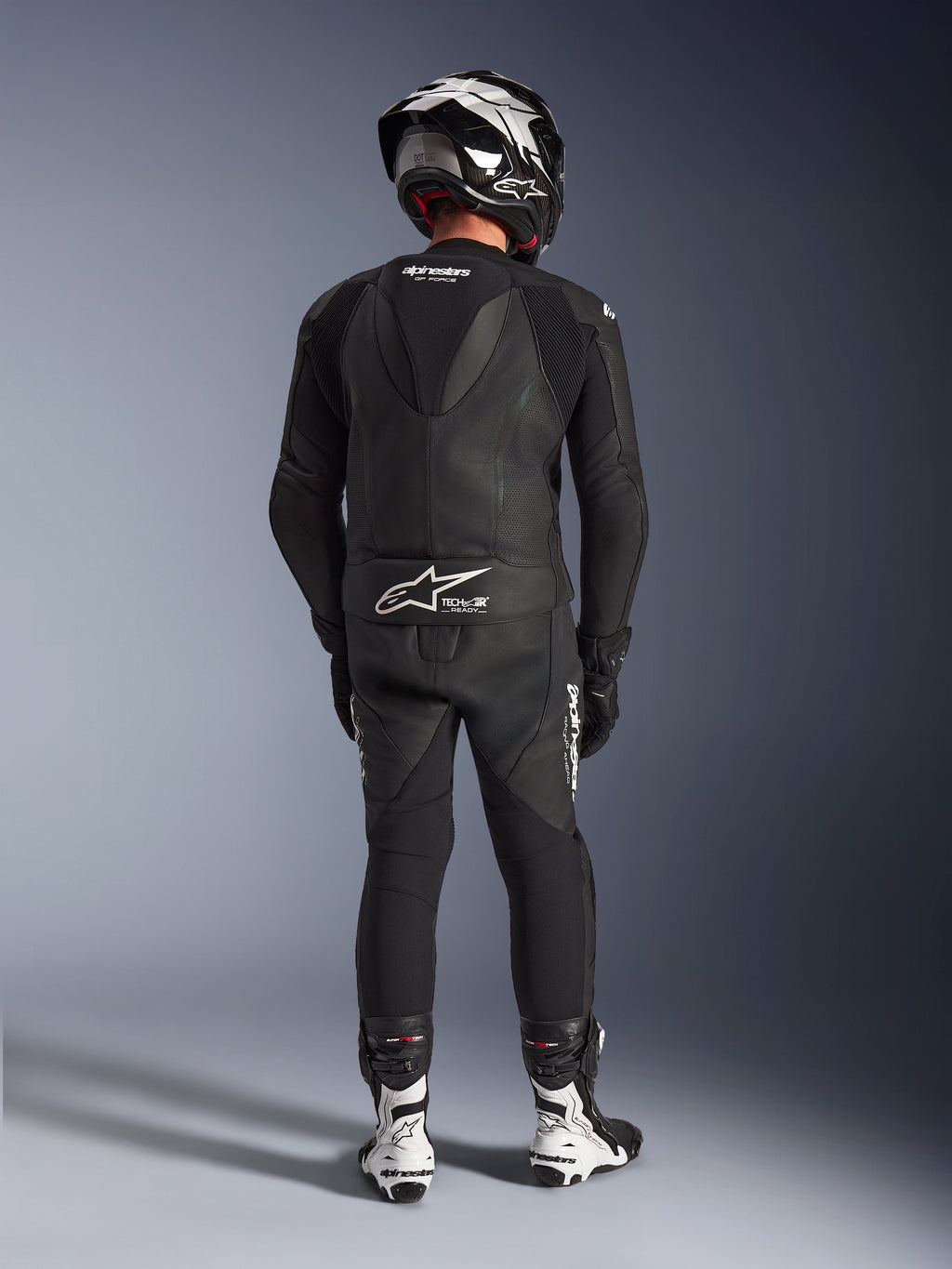 2-częściowy skórzany kombinezon Alpinestars GP Force V2 w kolorze czarnym, pełny widok z tyłu z widocznym aerodynamicznym garbem i brandingiem Tech-Air Ready, w zestawie z czarno-białym kaskiem i białymi butami wyścigowymi.