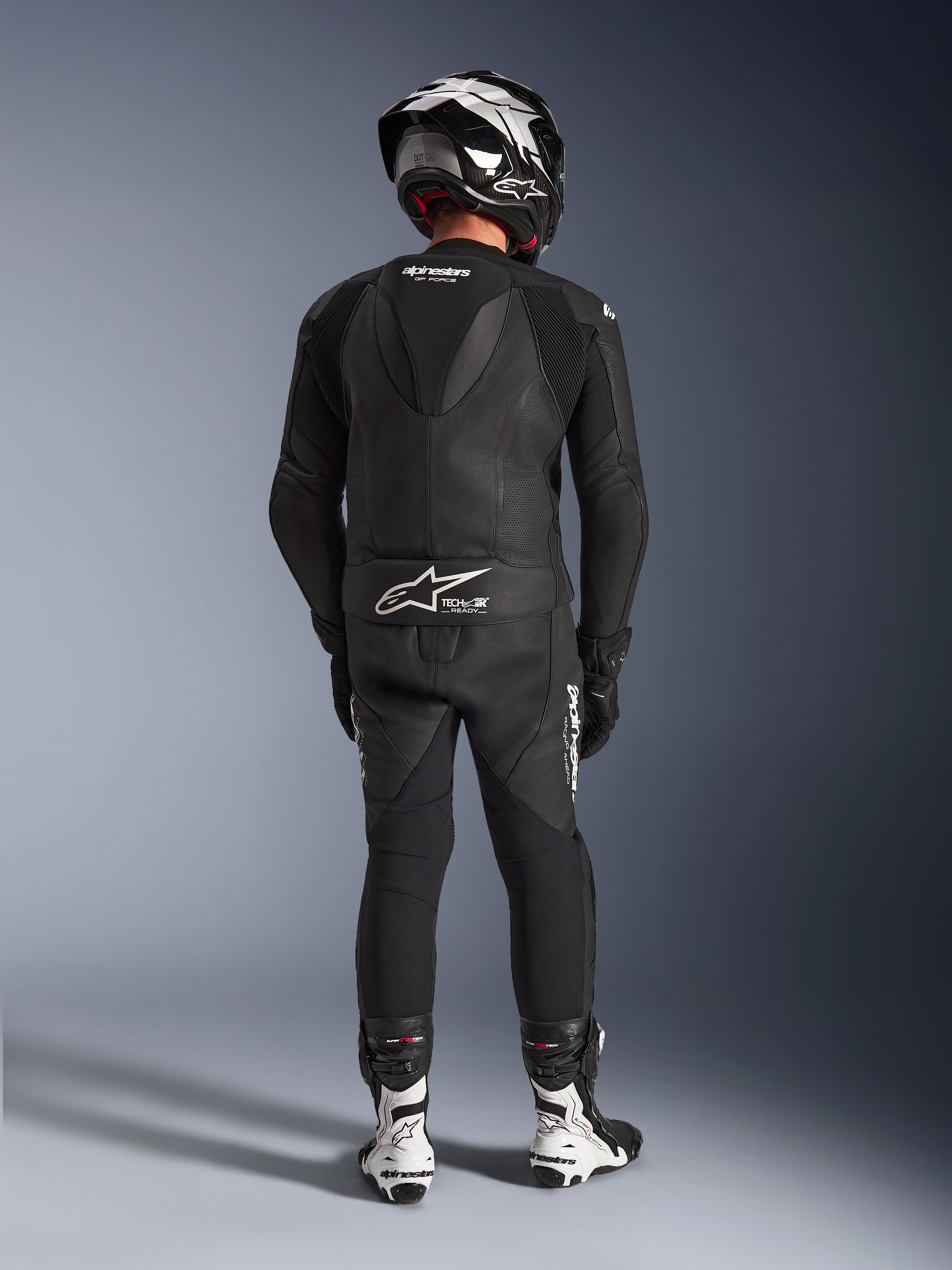 2-częściowy skórzany kombinezon Alpinestars GP Force V2 w kolorze czarnym, pełny widok z tyłu z widocznym aerodynamicznym garbem i brandingiem Tech-Air Ready, w zestawie z czarno-białym kaskiem i białymi butami wyścigowymi.