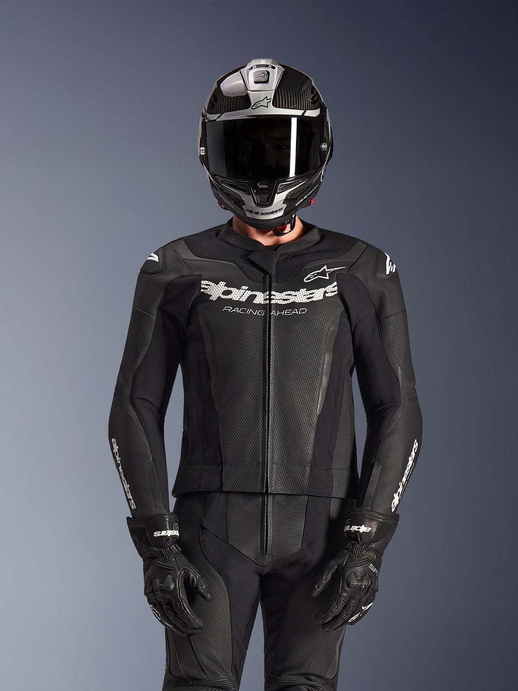 Motocyklista w dwuczęściowym skórzanym kombinezonie Alpinestars GP Force V2 w kolorze czarnym, konstrukcja z perforowanej skóry z białym logo „Racing Ahead” na klatce piersiowej, srebrny kask integralny z włókna węglowego, czarne rękawice, profesjonalna odzież sportowa i torowa