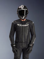 Motocyklista w dwuczęściowym skórzanym kombinezonie Alpinestars GP Force V2 w kolorze czarnym, konstrukcja z perforowanej skóry z białym logo „Racing Ahead” na klatce piersiowej, srebrny kask integralny z włókna węglowego, czarne rękawice, profesjonalna odzież sportowa i torowa