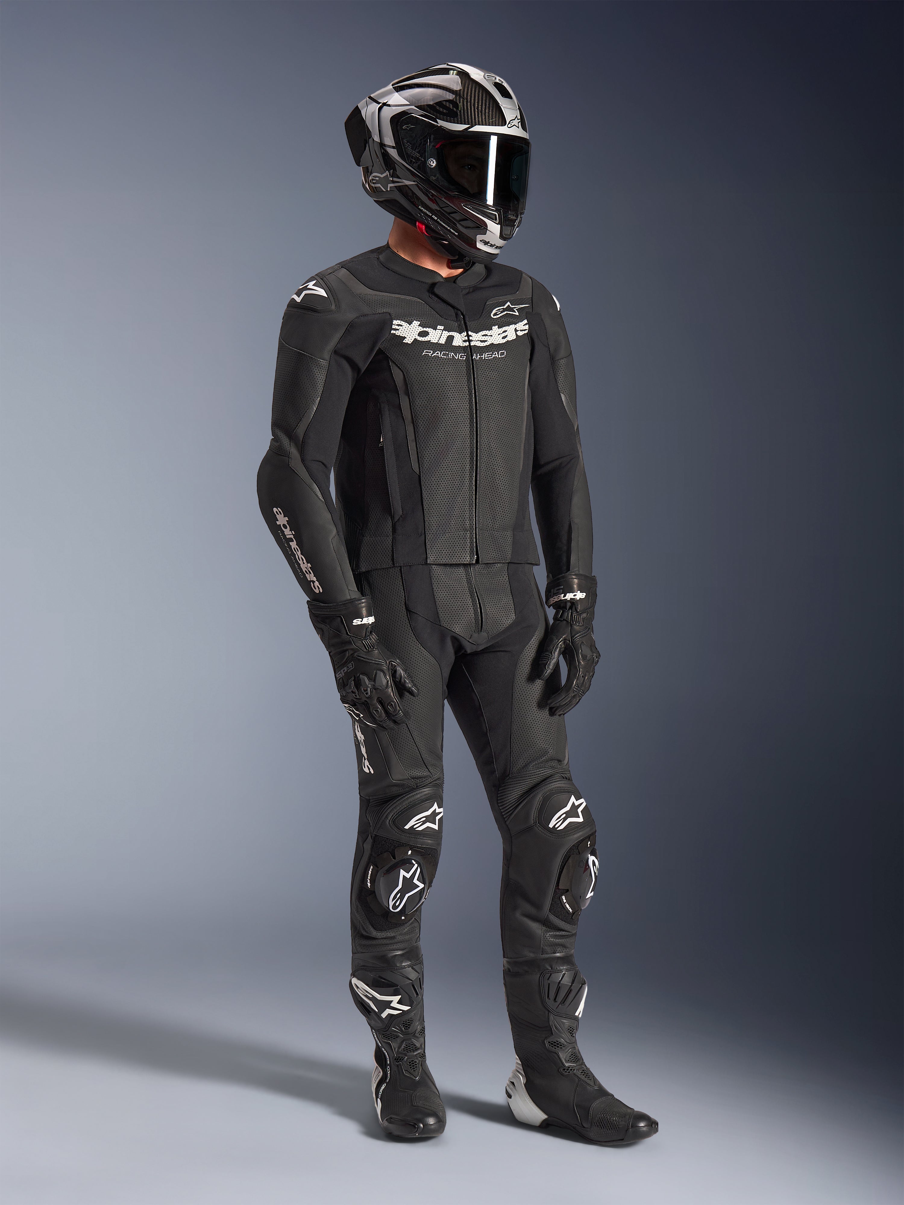 Alpinestars GP Force V2 2-Piece Leather Suit, czarny motocyklowy kombinezon wyścigowy, noszony z kaskiem integralnym, rękawicami i butami, wyposażony w perforowaną skórzaną konstrukcję, aerodynamiczny garb oraz białe logo Alpinestars na klatce piersiowej, ramionach i kolanach.
