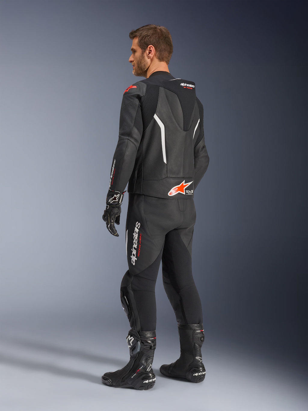 Alpinestars GP Force V2 2-Piece Leather Suit, dwuczęściowy kombinezon skórzany, czarny z białymi i czerwonymi akcentami fluo, widok z tyłu na modelu, aerodynamiczny garb, perforowane panele skórzane i wyraźne logo Alpinestars, czarne buty i rękawice wyścigowe.