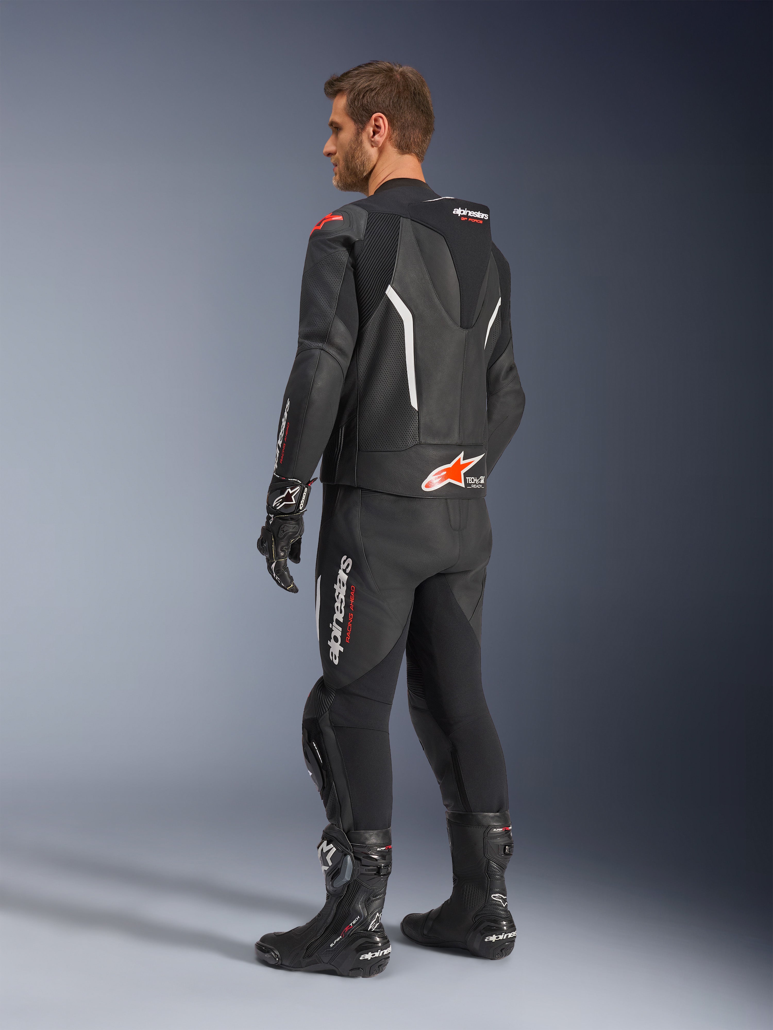Alpinestars GP Force V2 2-Piece Leather Suit, dwuczęściowy kombinezon skórzany, czarny z białymi i czerwonymi akcentami fluo, widok z tyłu na modelu, aerodynamiczny garb, perforowane panele skórzane i wyraźne logo Alpinestars, czarne buty i rękawice wyścigowe.