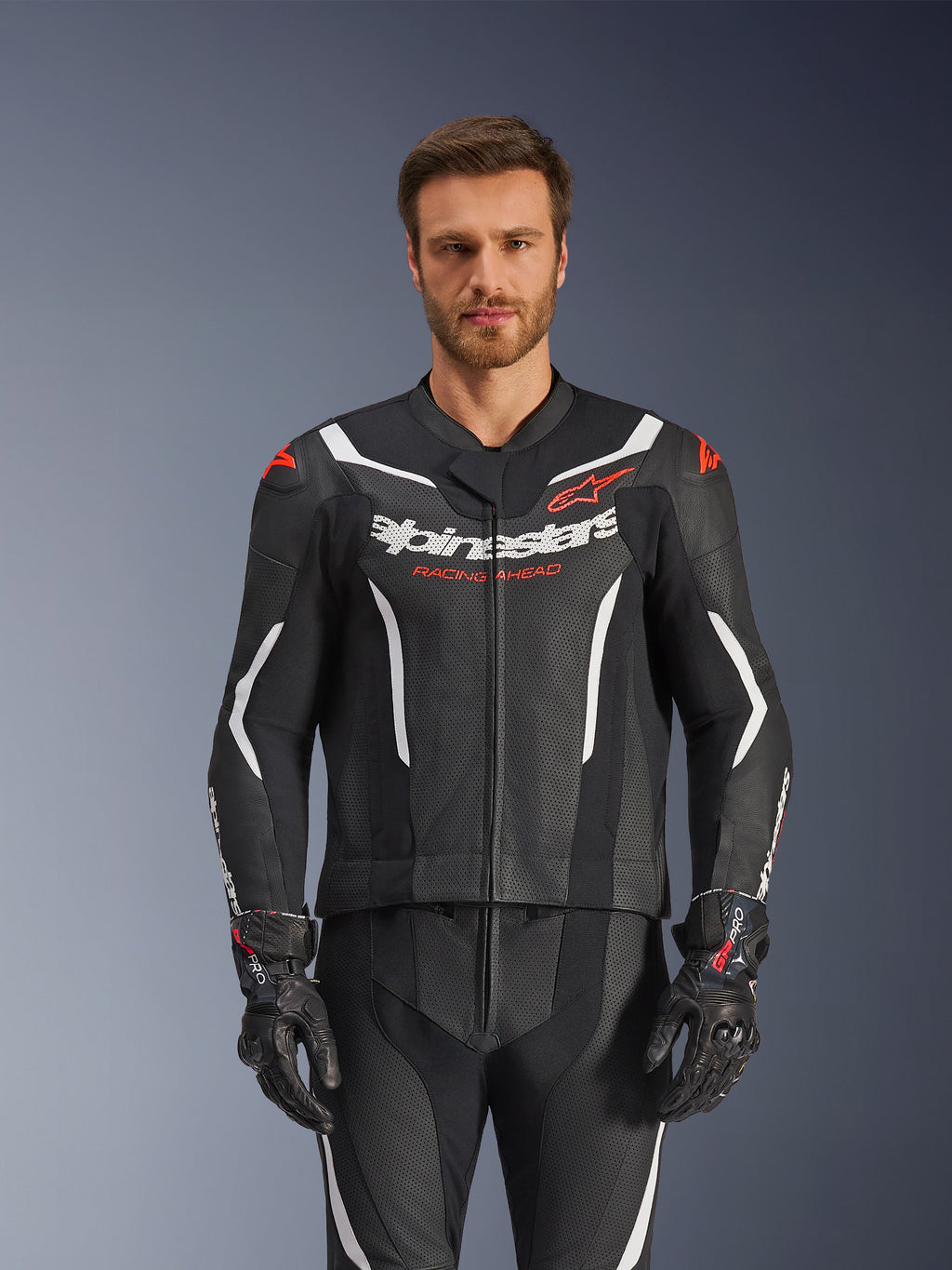 Alpinestars GP Force V2 Leather Suit- 2Piece, czarno-biało-czerwony fluo, widok z przodu na modela ubranego w perforowany kombinezon skórzany i pasujące czarne rękawice, z białymi kanciastymi akcentami i logo Alpinestars na klatce piersiowej