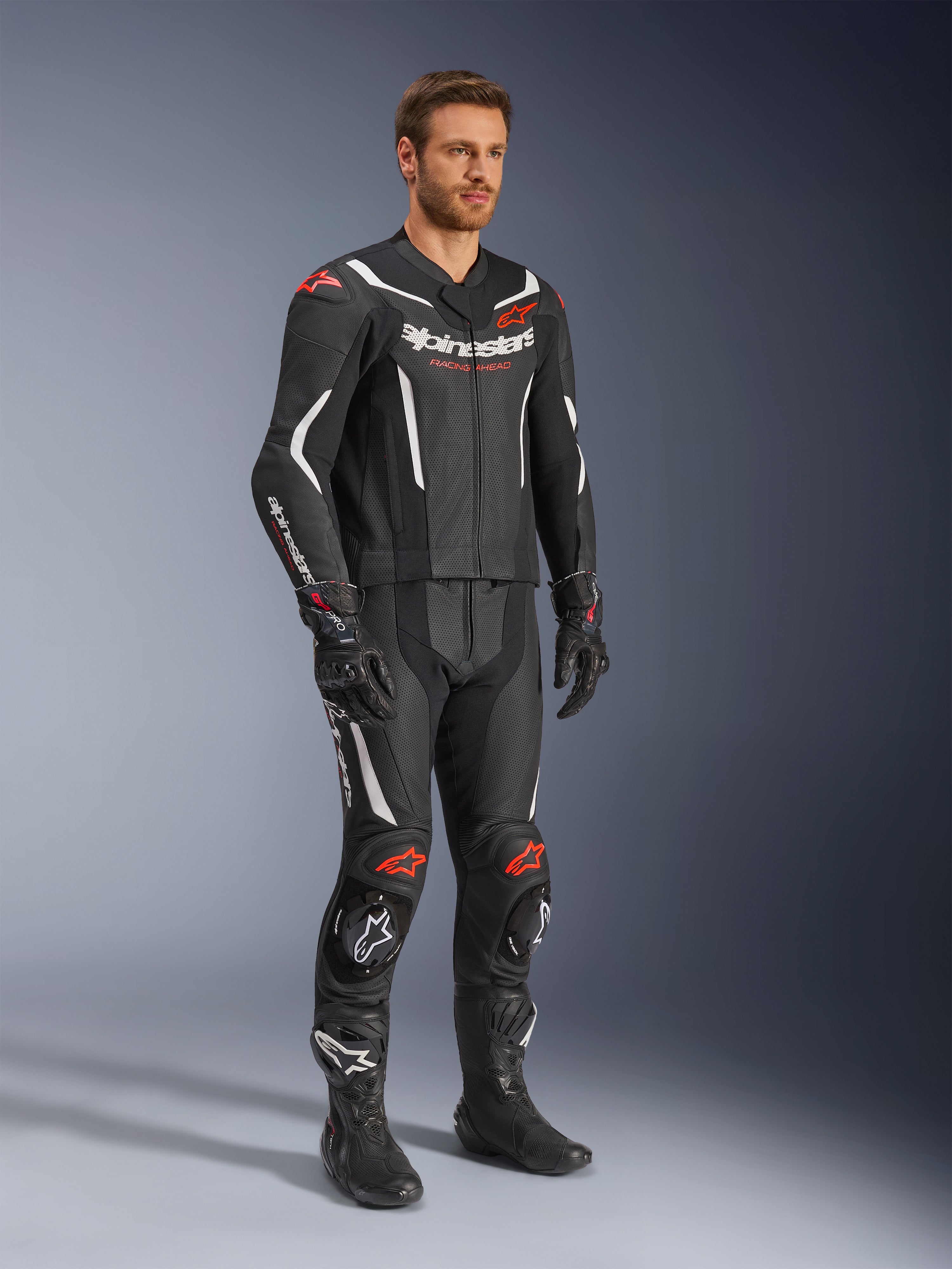 Alpinestars GP Force V2 2-Piece Leather Suit, czarny, biały i czerwony fluo, noszony przez modela z czarnymi butami i rękawicami wyścigowymi, z perforowanymi panelami skórzanymi, białymi akcentami aerodynamicznymi i logotypami w kolorze czerwonym fluo na ramionach i kolanach, profesjonalna odzież motocyklowa