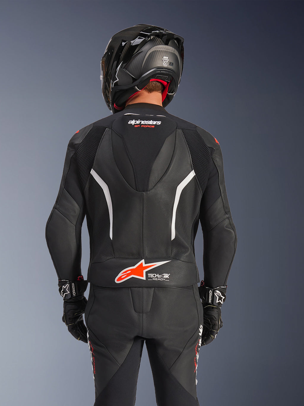 Alpinestars GP Force V2 2-Piece Leather Suit, widok z tyłu, czarny dwuczęściowy kombinezon skórzany z perforowaną skórą, białymi akcentami i czerwonym logo Alpinestars w dolnej części pleców, czarny kask motocyklowy i rękawice wyścigowe