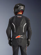 Alpinestars GP Force V2 2-Piece Leather Suit, widok z tyłu, czarny dwuczęściowy kombinezon skórzany z perforowaną skórą, białymi akcentami i czerwonym logo Alpinestars w dolnej części pleców, czarny kask motocyklowy i rękawice wyścigowe