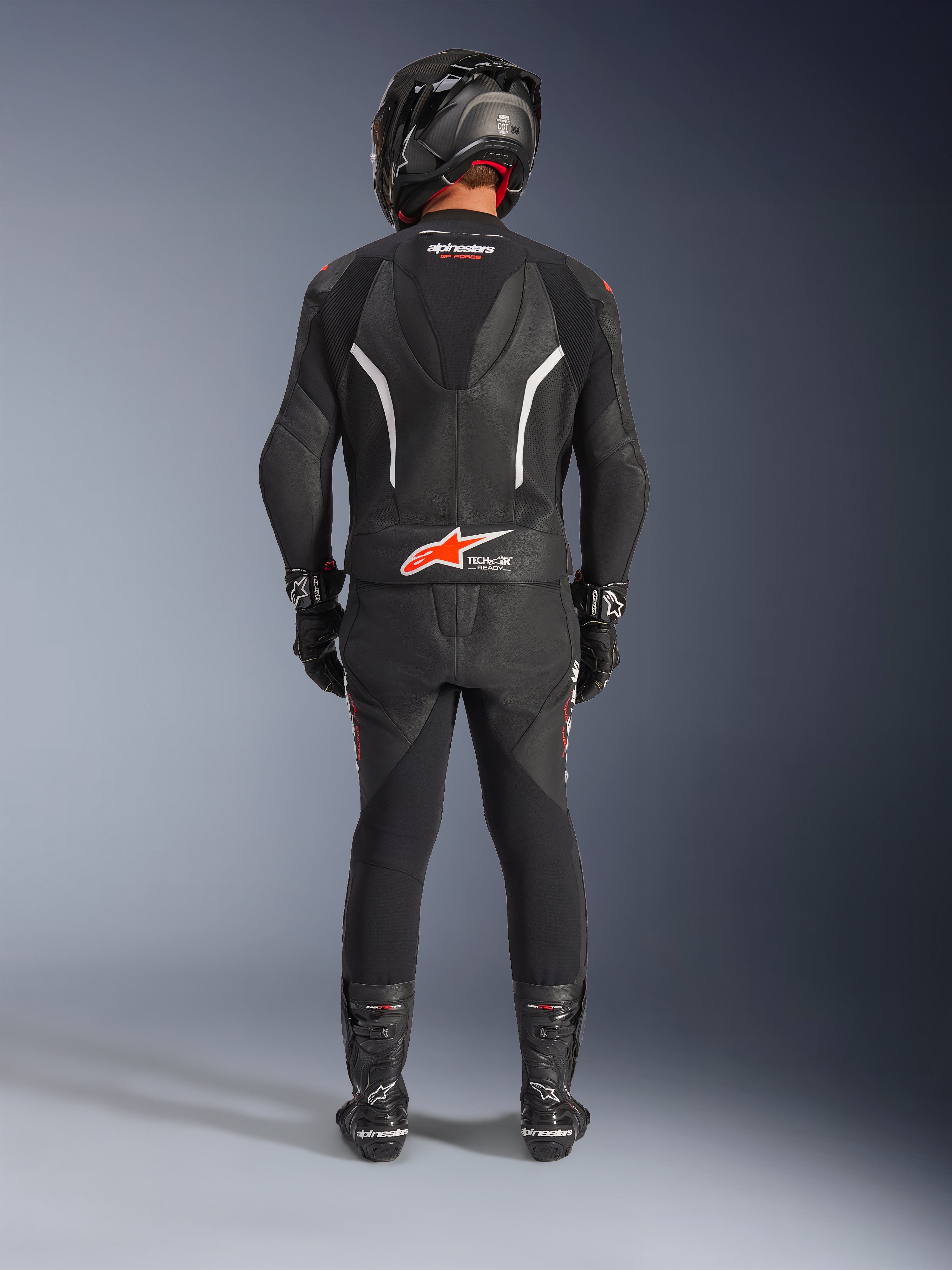 Motocyklista widziany od tyłu w dwuczęściowym skórzanym kombinezonie Alpinestars GP Force V2, czarny z białymi i czerwonymi akcentami fluo, z aerodynamicznym garbem i logo Tech-Air Ready, czarny kask wyścigowy, rękawice i buty.