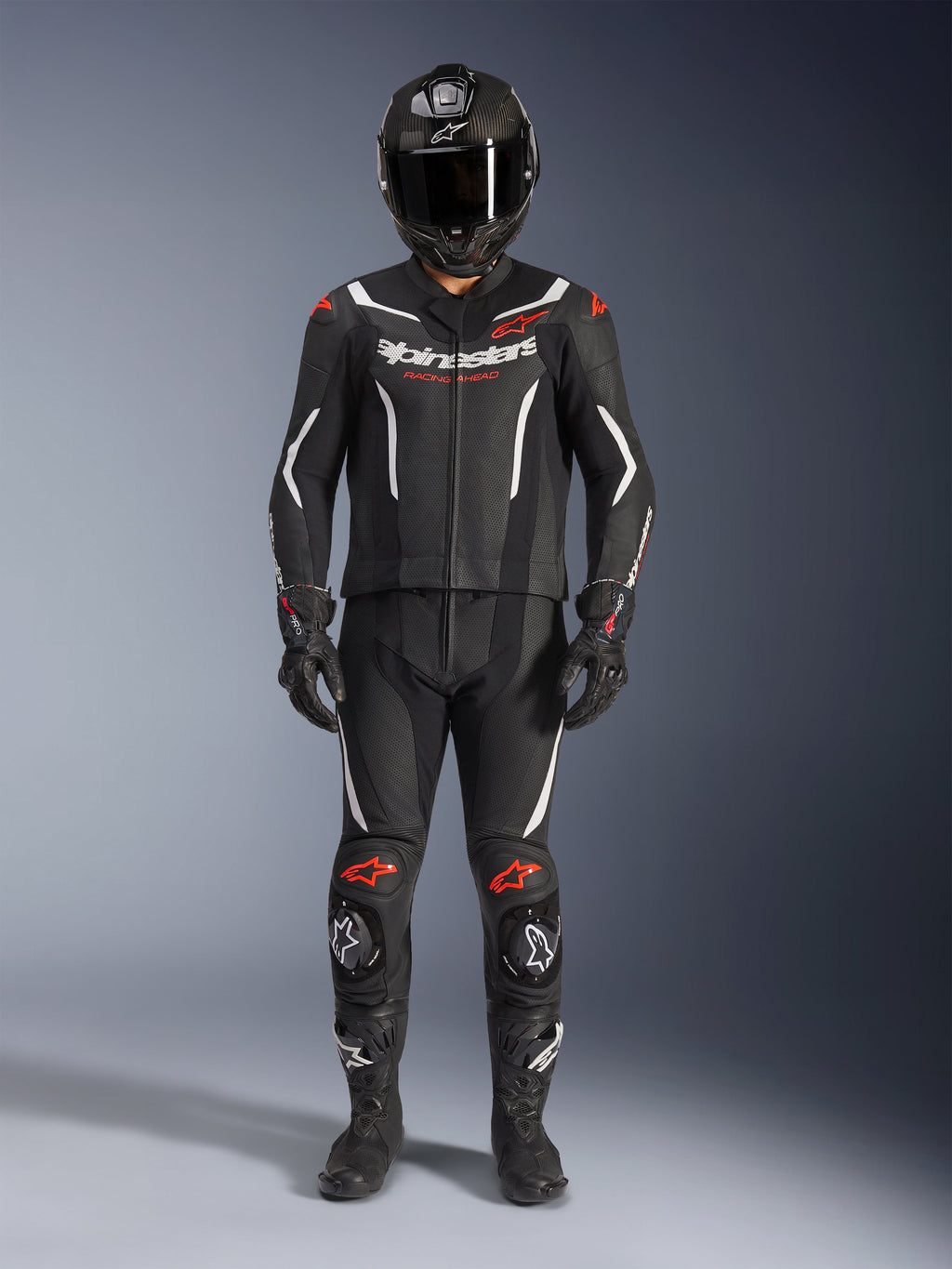 Alpinestars GP Force V2 Leather Suit 2-Piece, dwuczęściowy skórzany kombinezon motocyklowy, czarno-biały z akcentami Red Fluo, białe logo Alpinestars na klatce piersiowej, czerwone detale na ramionach i kolanach, w zestawie z czarnym kaskiem integralnym, czarnymi rękawicami i czarnymi butami.
