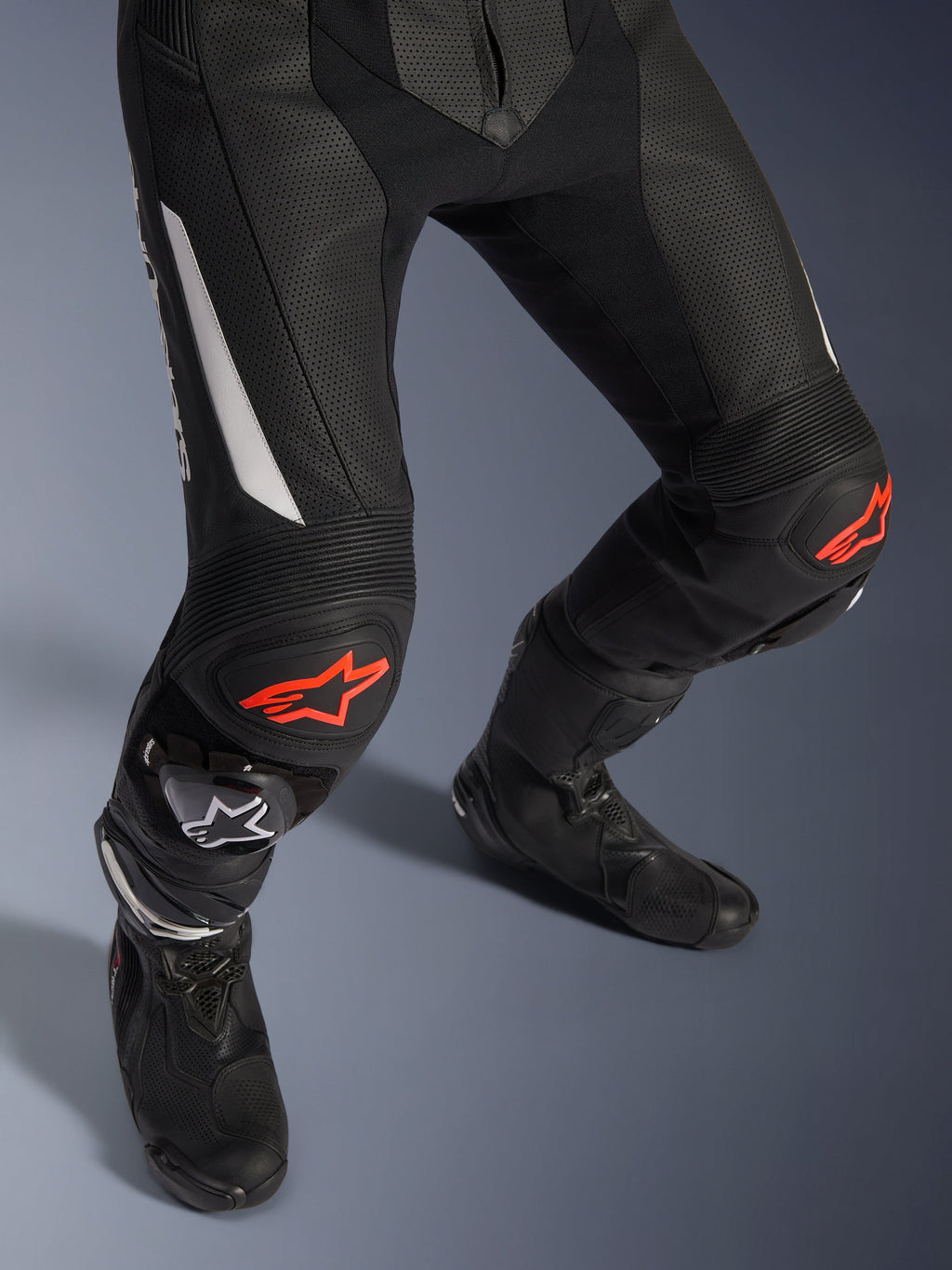 Dwuczęściowy skórzany kombinezon Alpinestars GP Force V2, motocyklowe spodnie wyścigowe w kolorze czarnym z czerwonymi i białymi akcentami, perforowana skóra, elastyczne panele harmonijkowe nad kolanami z czerwonym logo gwiazdy, czarne buty motocyklowe, wysokiej klasy odzież wyścigowa