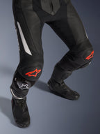 Dwuczęściowy skórzany kombinezon Alpinestars GP Force V2, motocyklowe spodnie wyścigowe w kolorze czarnym z czerwonymi i białymi akcentami, perforowana skóra, elastyczne panele harmonijkowe nad kolanami z czerwonym logo gwiazdy, czarne buty motocyklowe, wysokiej klasy odzież wyścigowa