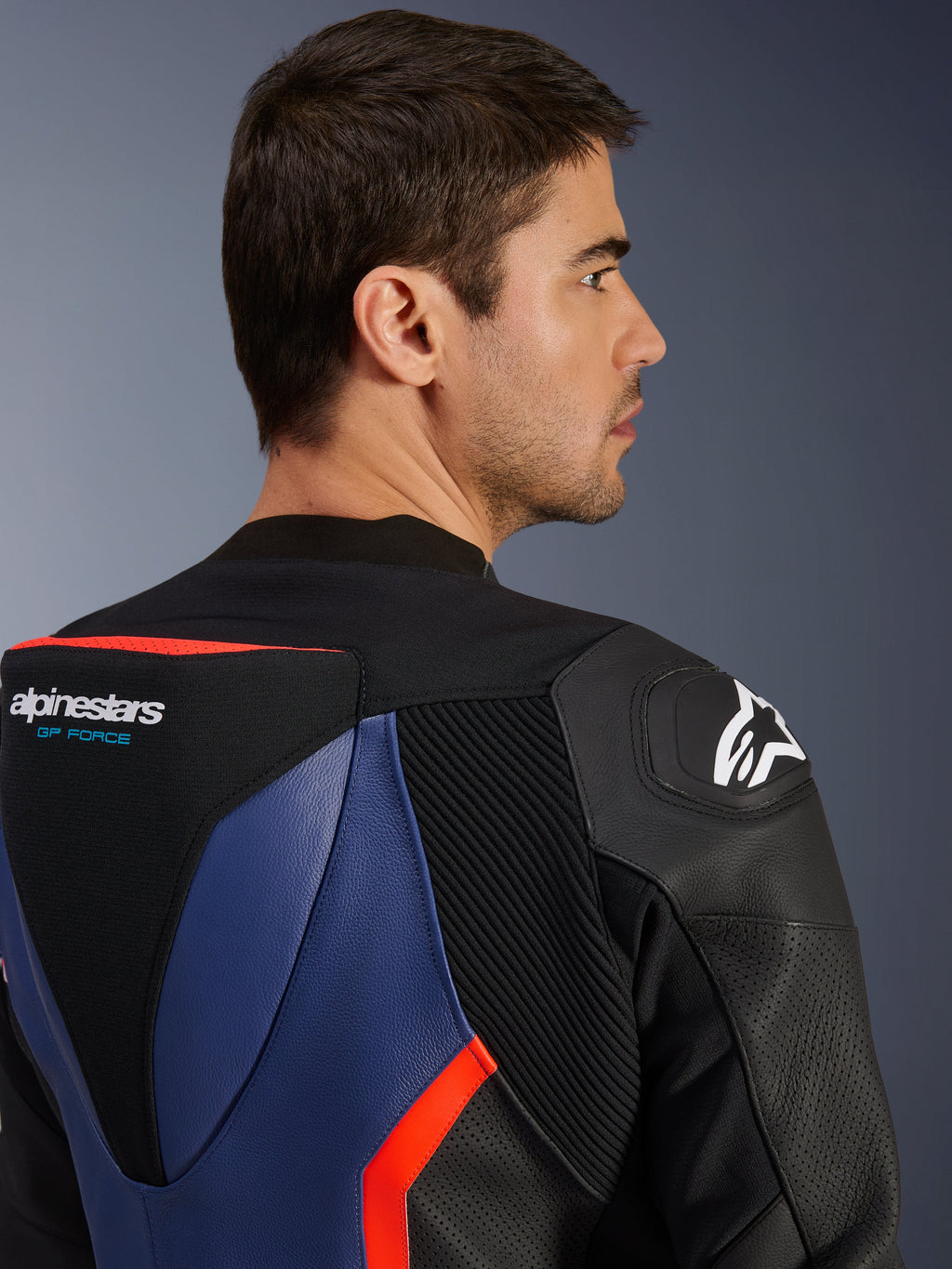 Dwuczęściowy kombinezon skórzany Alpinestars GP Force V2, zbliżenie na górną część pleców i ramię, garb aerodynamiczny z logo GP Force, slider ramion DFS, czarna, niebieska i czerwona fluo skóra bydlęca, perforowane panele i harmonijkowe strefy elastyczne, profesjonalna odzież wyścigowa