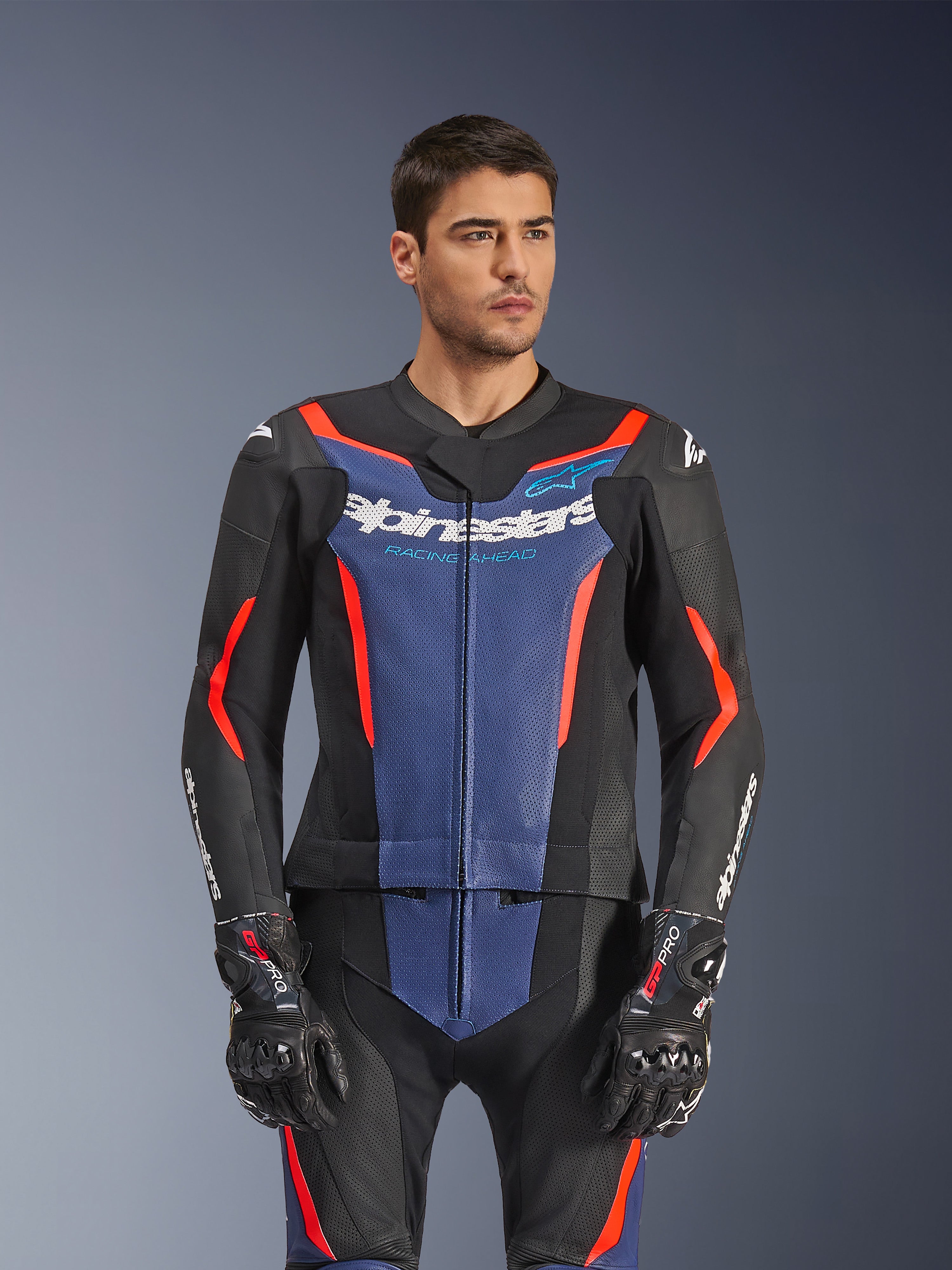Męski model w dwuczęściowym skórzanym kombinezonie Alpinestars GP Force V2, kolor Black Blue Red Fluo, z wyraźnym niebieskim panelem na klatce piersiowej i czerwonymi akcentami, zestawiony z rękawicami GP Pro, profesjonalna skórzana odzież motocyklowa o perforowanej teksturze