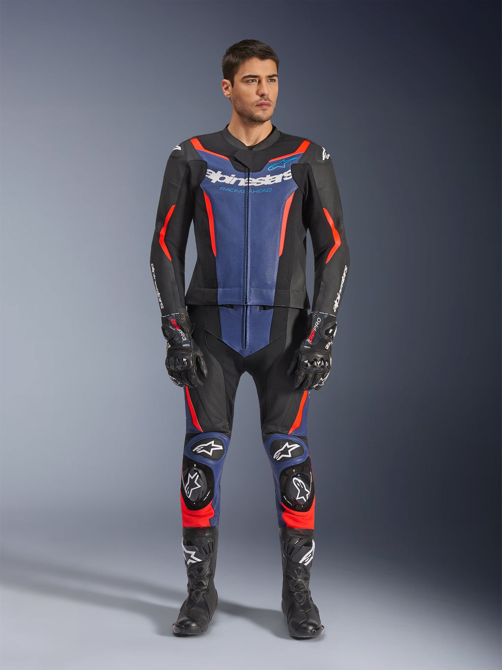 Alpinestars GP Force V2 Leather Suit 2-Piece, dwuczęściowy kombinezon skórzany w kolorze czarnym, niebieskim i czerwonym fluo, niebieskie panele na klatce piersiowej i dolnej części nogawek z jaskrawymi czerwonymi akcentami, prezentowany przez modela w czarnych butach i rękawicach wyścigowych, profesjonalna odzież ochronna do sportów motorowych