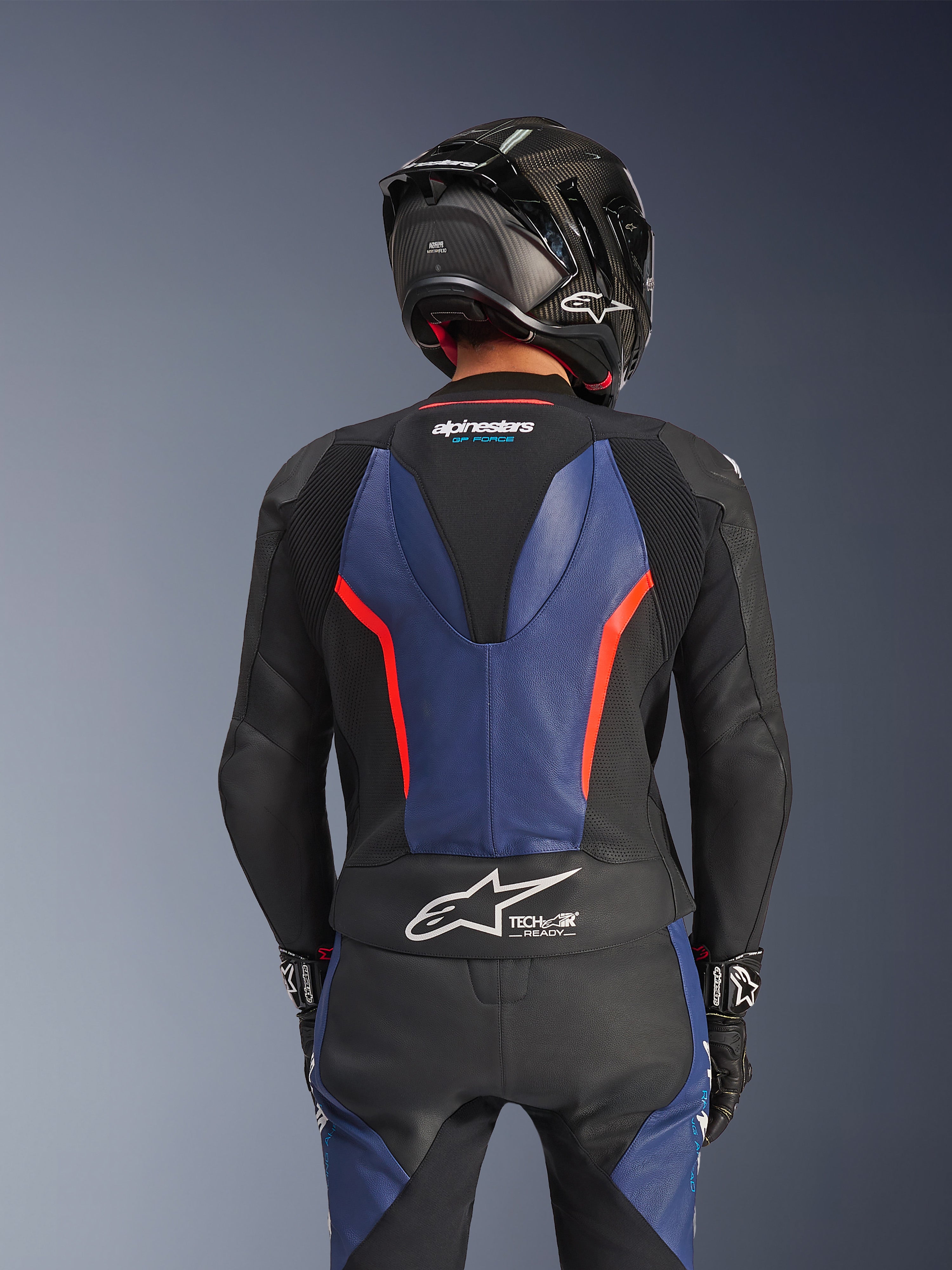 Dwuczęściowy skórzany kombinezon Alpinestars GP Force V2, kolor czarny, niebieski i neonowy czerwony, widok z tyłu ukazujący aerodynamiczny garb oraz oznaczenie Tech-Air Ready, najwyższej jakości skóra bydlęca ze strefami elastycznymi, czarny kask z włókna węglowego, profesjonalny sprzęt motocyklowy