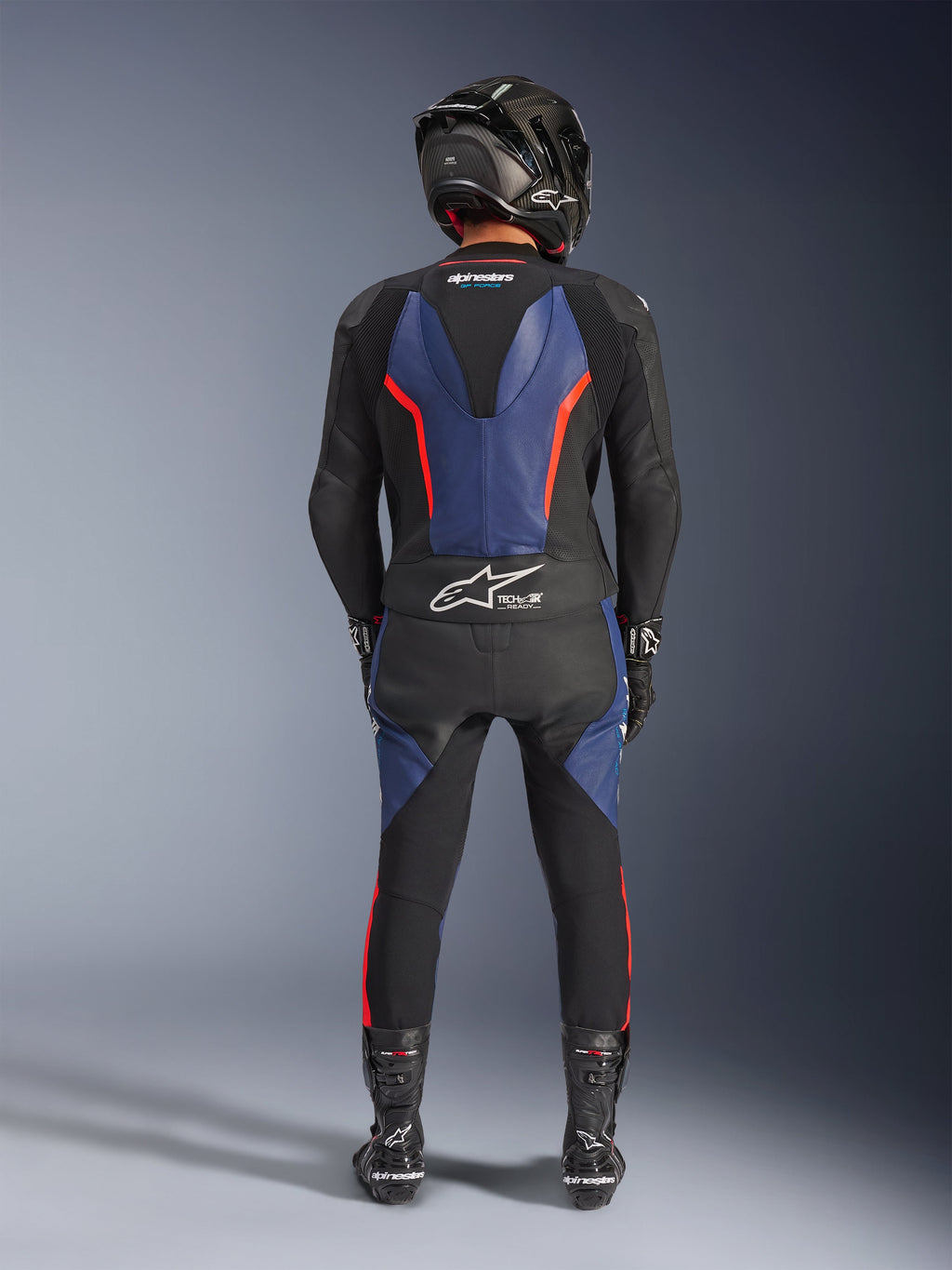 Alpinestars GP Force V2 Leather Suit 2-Piece, Black Blue Red Fluo, pełny widok z tyłu na motocyklistę w pełnym stroju wyścigowym, niebieskie panele skórzane na plecach i nogach z fluorescencyjnymi czerwonymi akcentami, logo Tech-Air® Ready na dolnej części pleców, kask z włókna węglowego, czarne rękawice i buty wyścigowe.