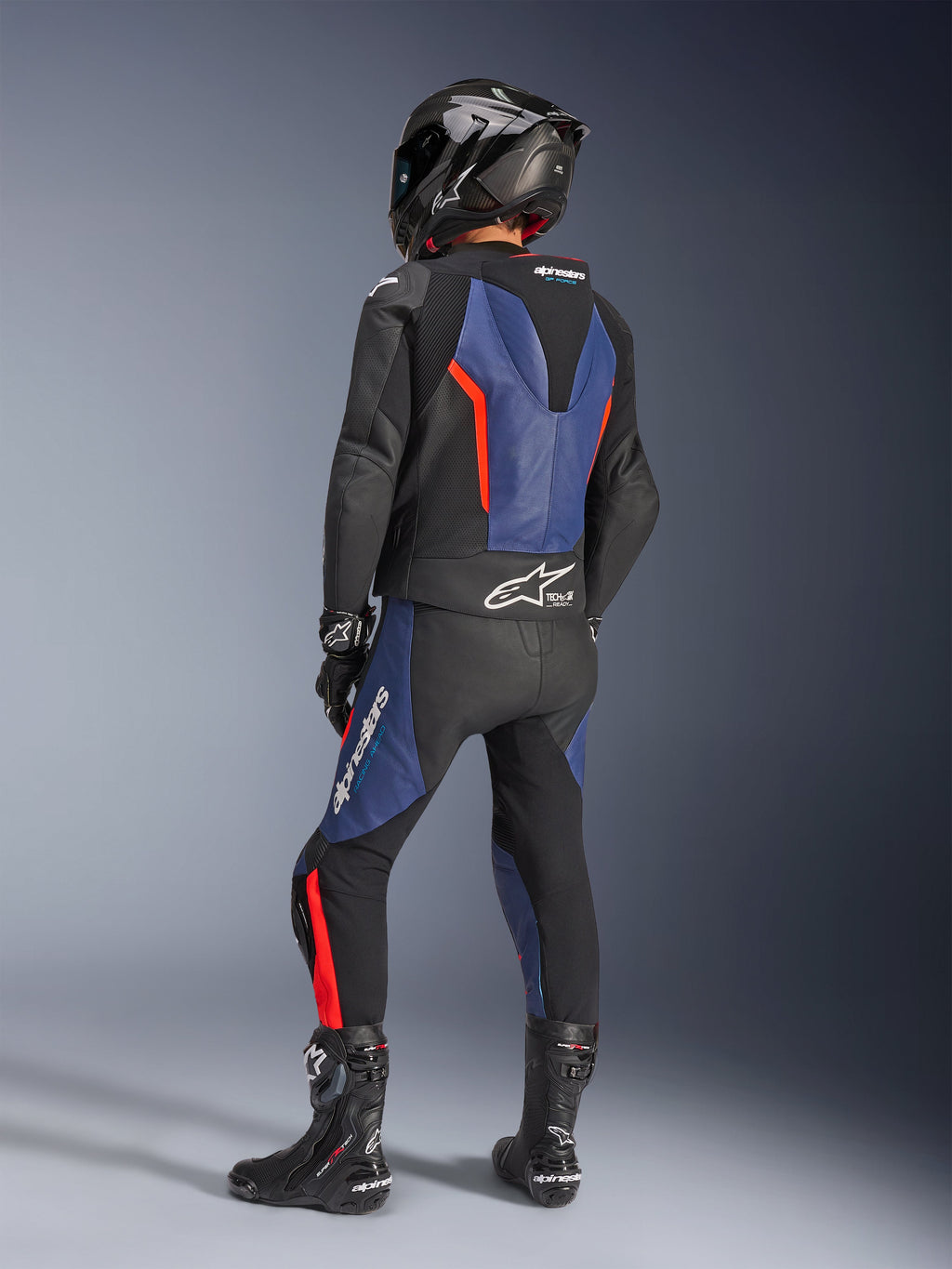 Alpinestars GP Force V2 Leather Suit 2-Piece, czarno-niebiesko-czerwony fluo, widok z tyłu motocyklisty w profesjonalnym stroju wyścigowym, zintegrowany garb aerodynamiczny i logo Tech-Air Ready, niebieskie i czerwone akcenty fluo na czarnej skórze, kask z włókna węglowego i czarne buty, wysokiej klasy odzież motocyklowa