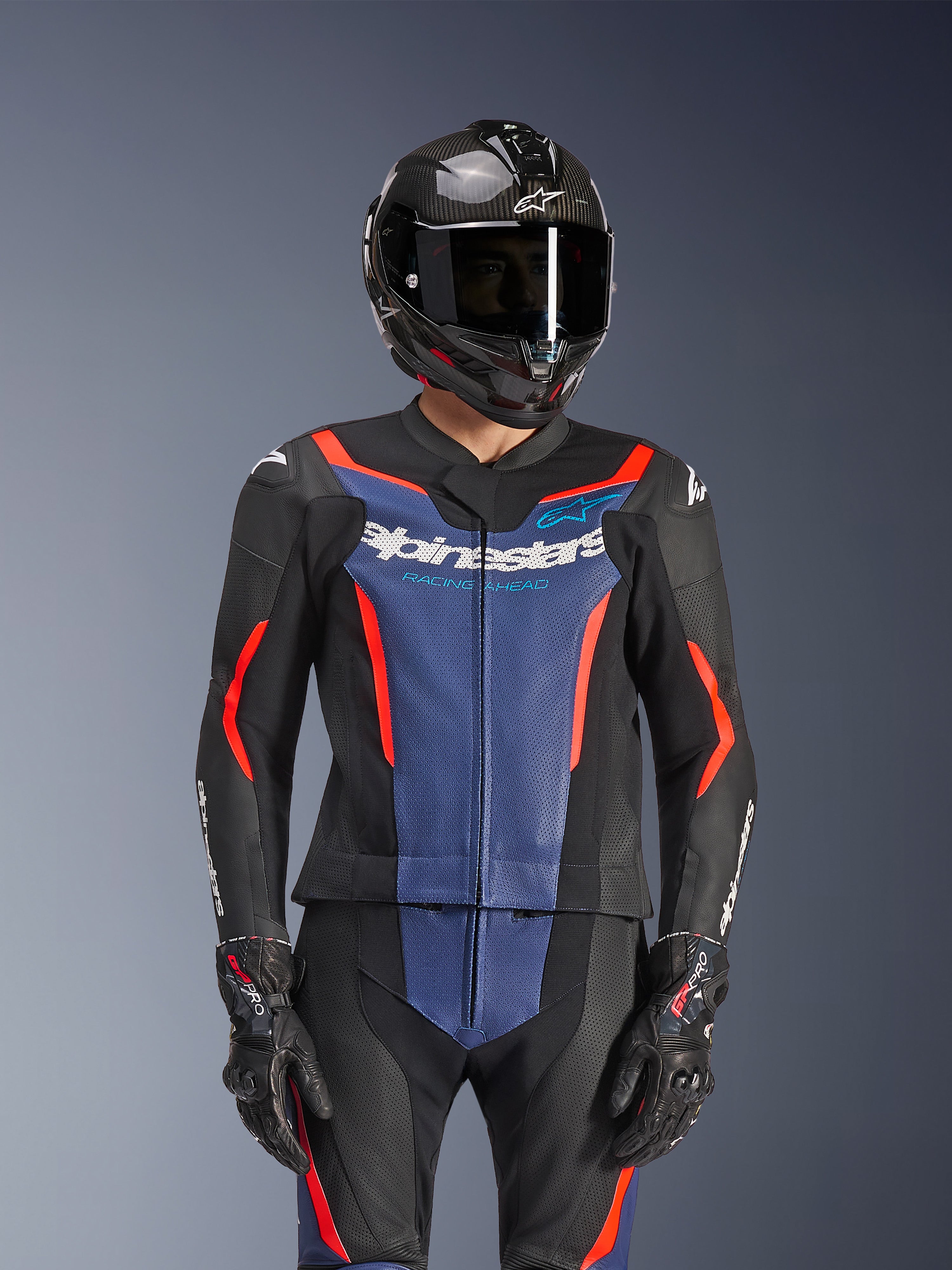 Alpinestars GP Force V2 Leather Suit 2-Piece, czarny, niebieski i czerwony fluo, noszony z kaskiem z włókna węglowego i rękawicami wyścigowymi, posiada perforowane panele skórzane i białe logo Alpinestars na klatce piersiowej, profesjonalna odzież do jazdy wyścigowej i sportowej
