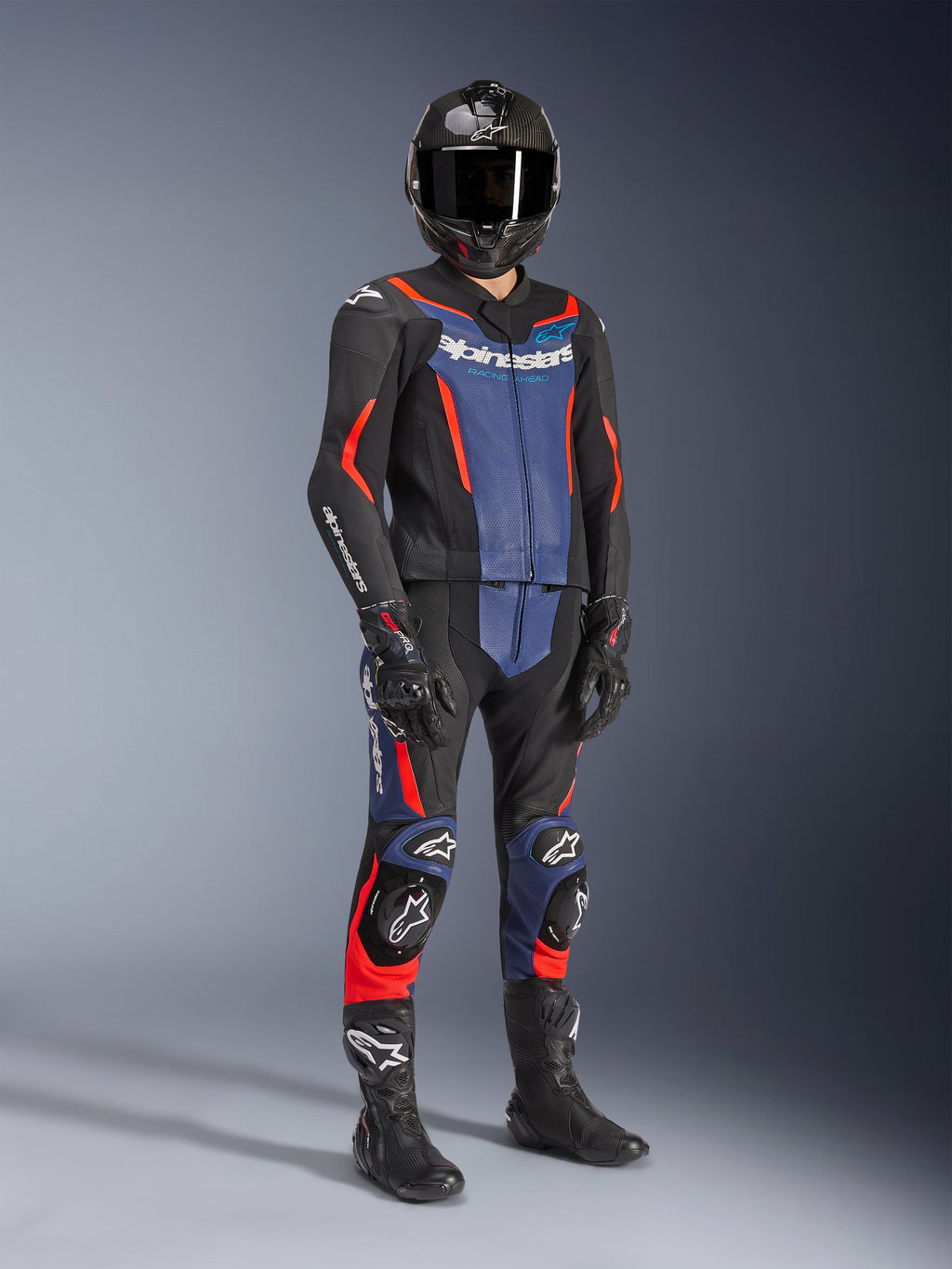 Dwuczęściowy skórzany kombinezon Alpinestars GP Force V2 w kolorze czarnym, niebieskim i czerwonym fluo, noszony z kaskiem z włókna węglowego, czarnymi rękawicami wyścigowymi i butami, z perforowanymi panelami skórzanymi i białym logo Alpinestars, profesjonalna sportowa odzież motocyklowa.