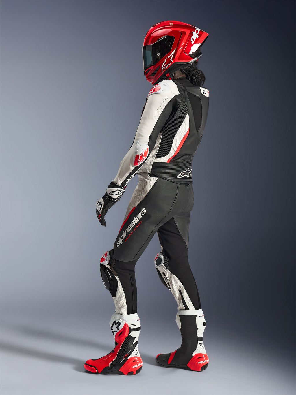 Motocyklistka w dwuczęściowym skórzanym kombinezonie Alpinestars Stella GP-R7, czarno-biało-czerwony fluo, widok z tyłu pod kątem prezentujący garb aerodynamiczny, czerwony kask integralny, czarno-białe rękawice wyścigowe i czerwono-białe techniczne buty wyścigowe, profesjonalna odzież motocyklowa