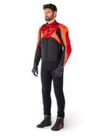 Męski motocyklista w kurtce Alpinestars T-Jaws V4 Waterproof w kolorze Black Red Fluo Dark Red, w zestawieniu z czarnymi spodniami motocyklowymi, rękawicami i butami do jazdy, o sportowym tekstylnym kroju z wyraźnym logo na klatce piersiowej i agresywnej sylwetce.