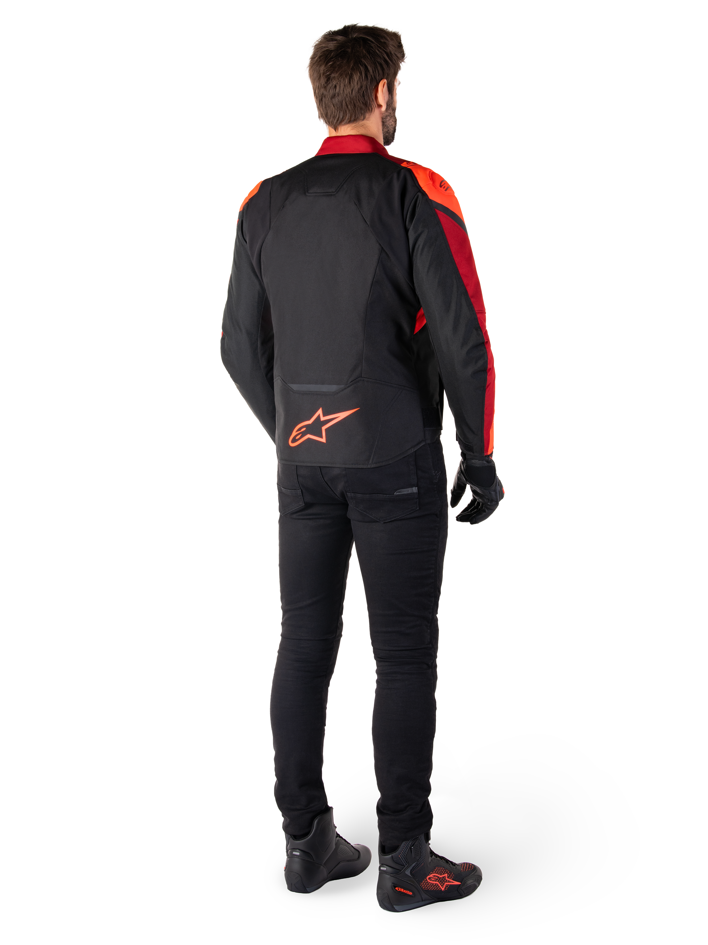 Alpinestars T-Jaws V4 Waterproof Jacket w kolorze Black Red Fluo Dark Red, widok z tyłu motocyklisty ubranego w kurtkę tekstylną, czarne spodnie i buty motocyklowe, z wyraźnym czerwonym logo Alpinestars w dolnej części pleców.