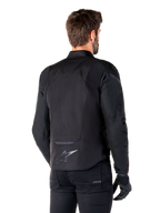 Kurtka wodoodporna Alpinestars T-Jaws V4, czarna, widok z tyłu na modelu, odblaskowe logo Alpinestars w dolnej części pleców, przedłużony tył dla lepszej osłony, sportowy tekstylny design z tonalnymi panelami, noszona z czarnymi rękawicami motocyklowymi