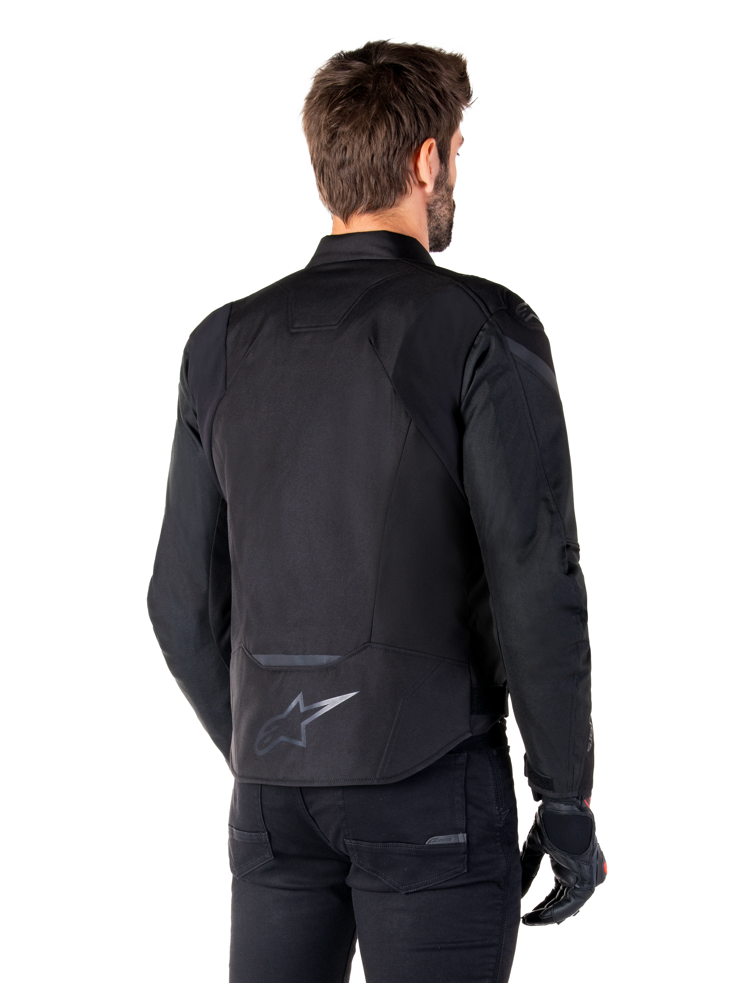 Kurtka wodoodporna Alpinestars T-Jaws V4, czarna, widok z tyłu na modelu, odblaskowe logo Alpinestars w dolnej części pleców, przedłużony tył dla lepszej osłony, sportowy tekstylny design z tonalnymi panelami, noszona z czarnymi rękawicami motocyklowymi