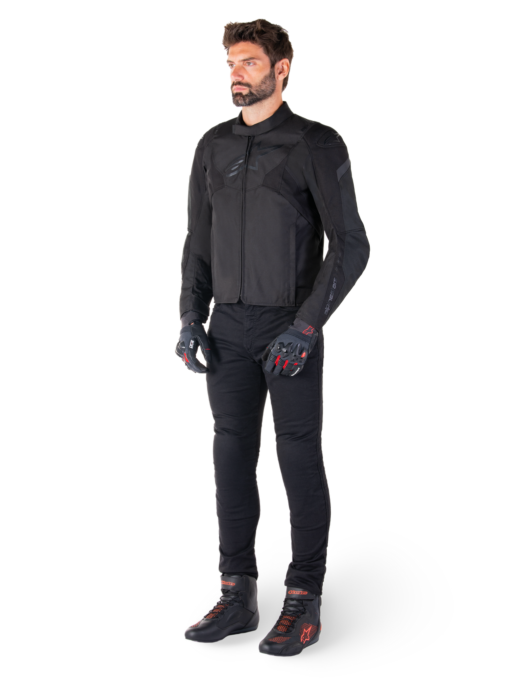 Męski model ubrany w wodoodporną kurtkę Alpinestars T-Jaws V4 w kolorze czarnym, w zestawieniu z czarnymi spodniami motocyklowymi, czarnymi rękawicami z czerwonymi akcentami oraz czarnymi butami motocyklowymi; kurtka o dopasowanym sportowym kroju z subtelnym brandingiem, na białym tle.