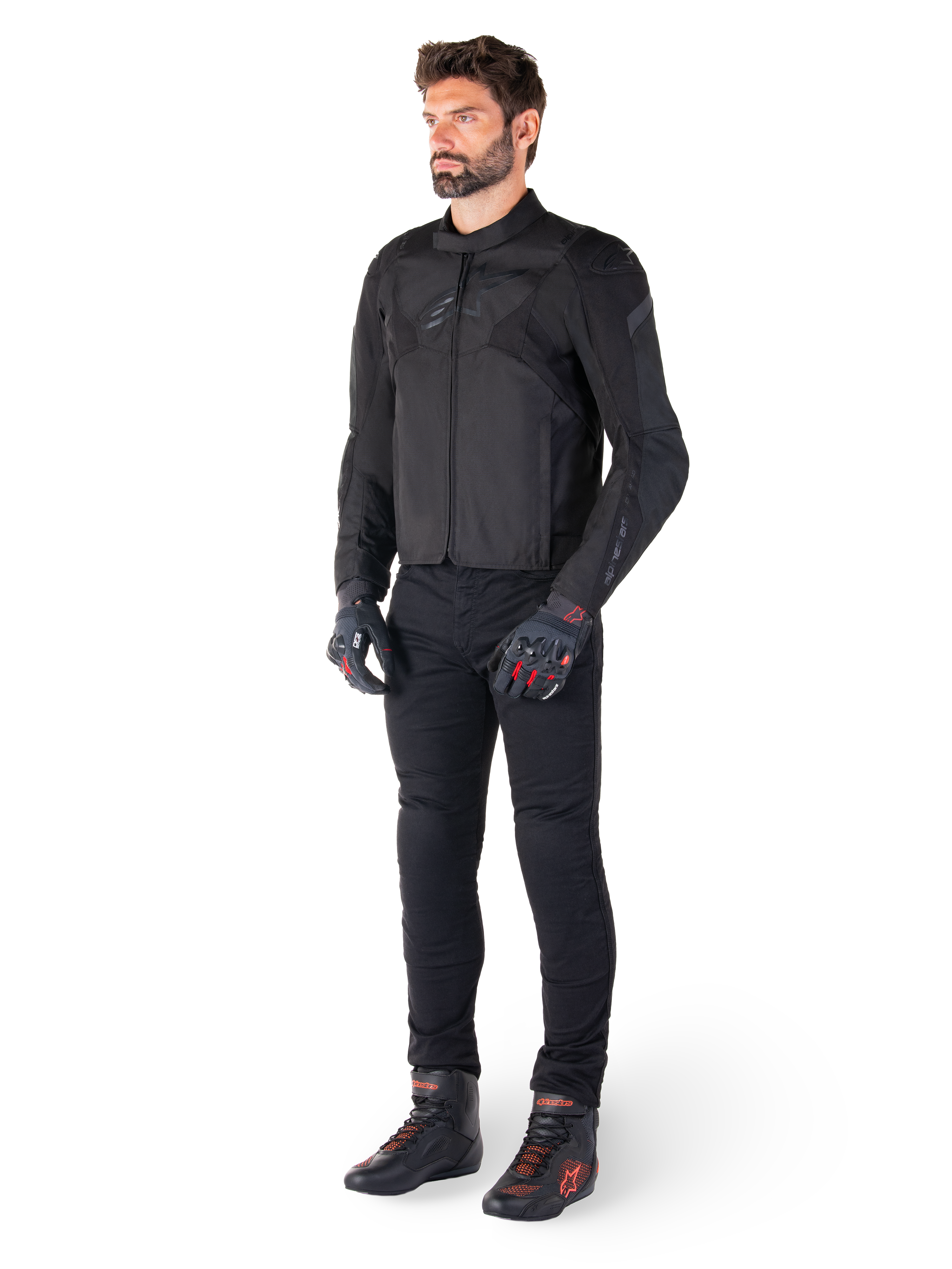 Męski model ubrany w wodoodporną kurtkę Alpinestars T-Jaws V4 w kolorze czarnym, w zestawieniu z czarnymi spodniami motocyklowymi, czarnymi rękawicami z czerwonymi akcentami oraz czarnymi butami motocyklowymi; kurtka o dopasowanym sportowym kroju z subtelnym brandingiem, na białym tle.