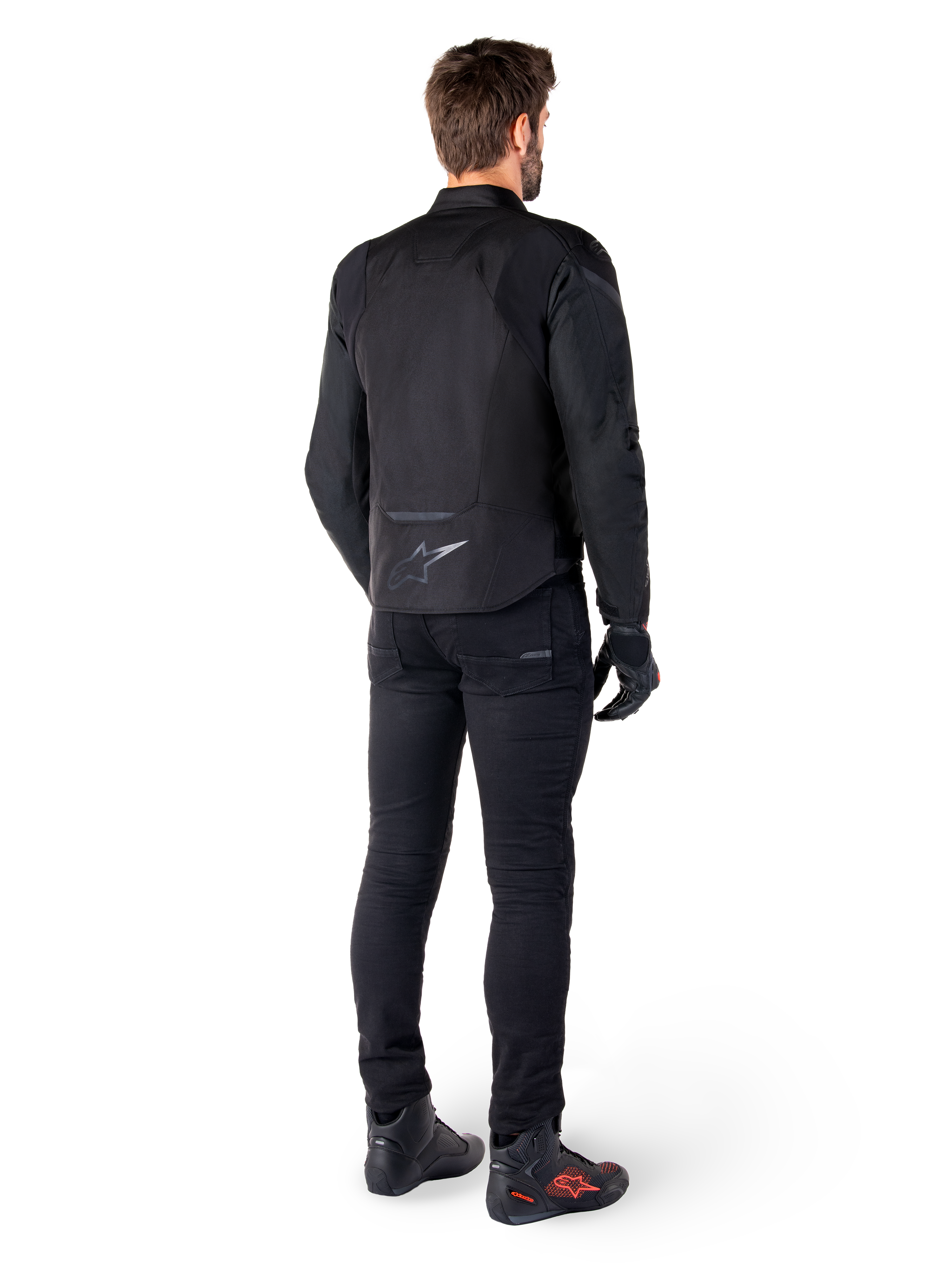 Alpinestars T-Jaws V4 Waterproof Jacket, czarna, widok z tyłu na modelu w czarnych spodniach i butach motocyklowych, sportowa konstrukcja tekstylna, logo Alpinestars w dolnej części pleców