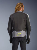 Wodoodporna kurtka Alpinestars T-Jaws V4, Black Ice Gray Dark Gray, widok z tyłu na modelu, jasnoszary panel w dolnej części pleców z wyraźnym neonowożółtym logo gwiazdy, wielopanelowa konstrukcja tekstylna, sportowa sylwetka, czarne rękawice i ciemne spodnie