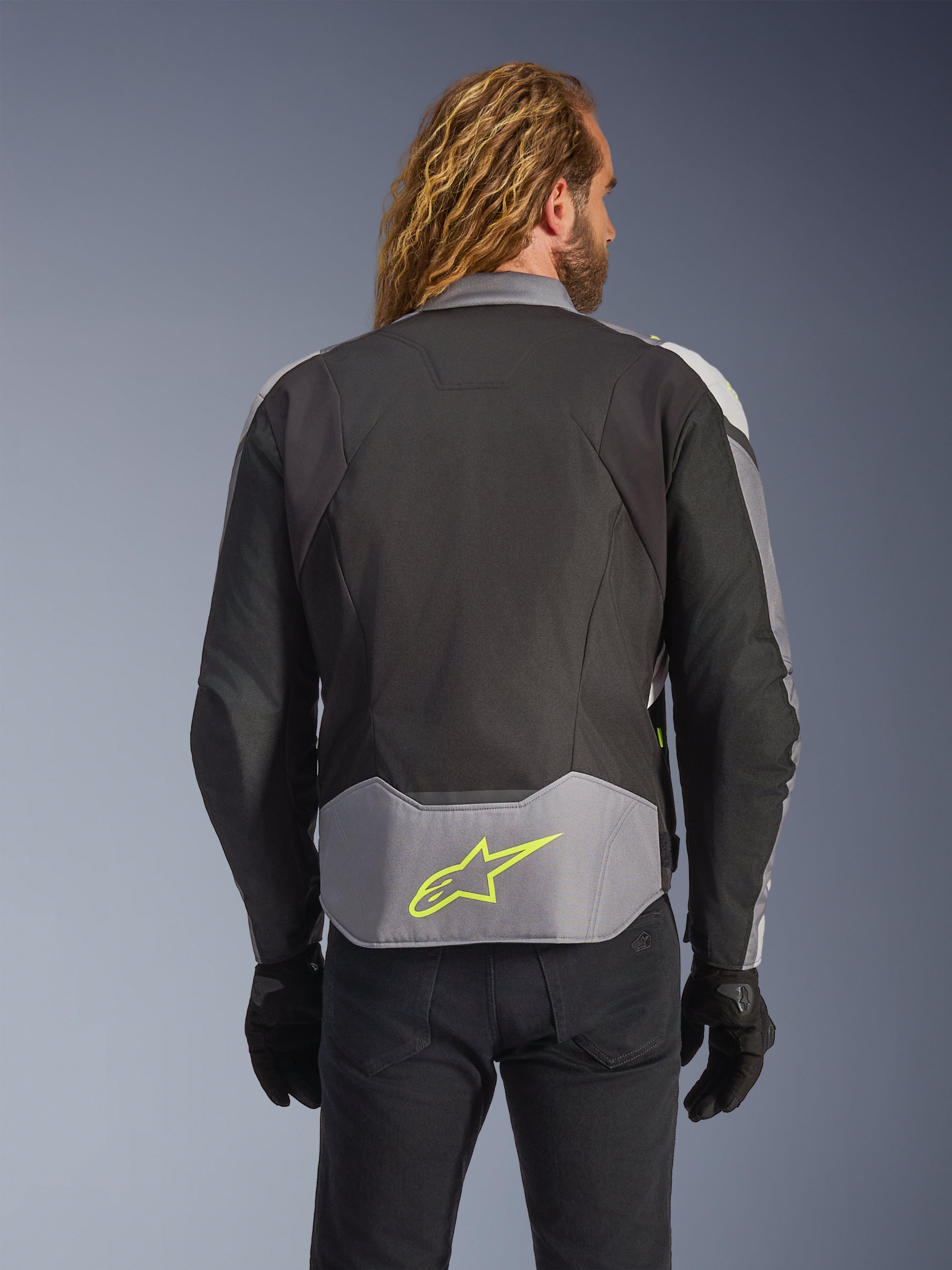 Wodoodporna kurtka Alpinestars T-Jaws V4, Black Ice Gray Dark Gray, widok z tyłu na modelu, jasnoszary panel w dolnej części pleców z wyraźnym neonowożółtym logo gwiazdy, wielopanelowa konstrukcja tekstylna, sportowa sylwetka, czarne rękawice i ciemne spodnie